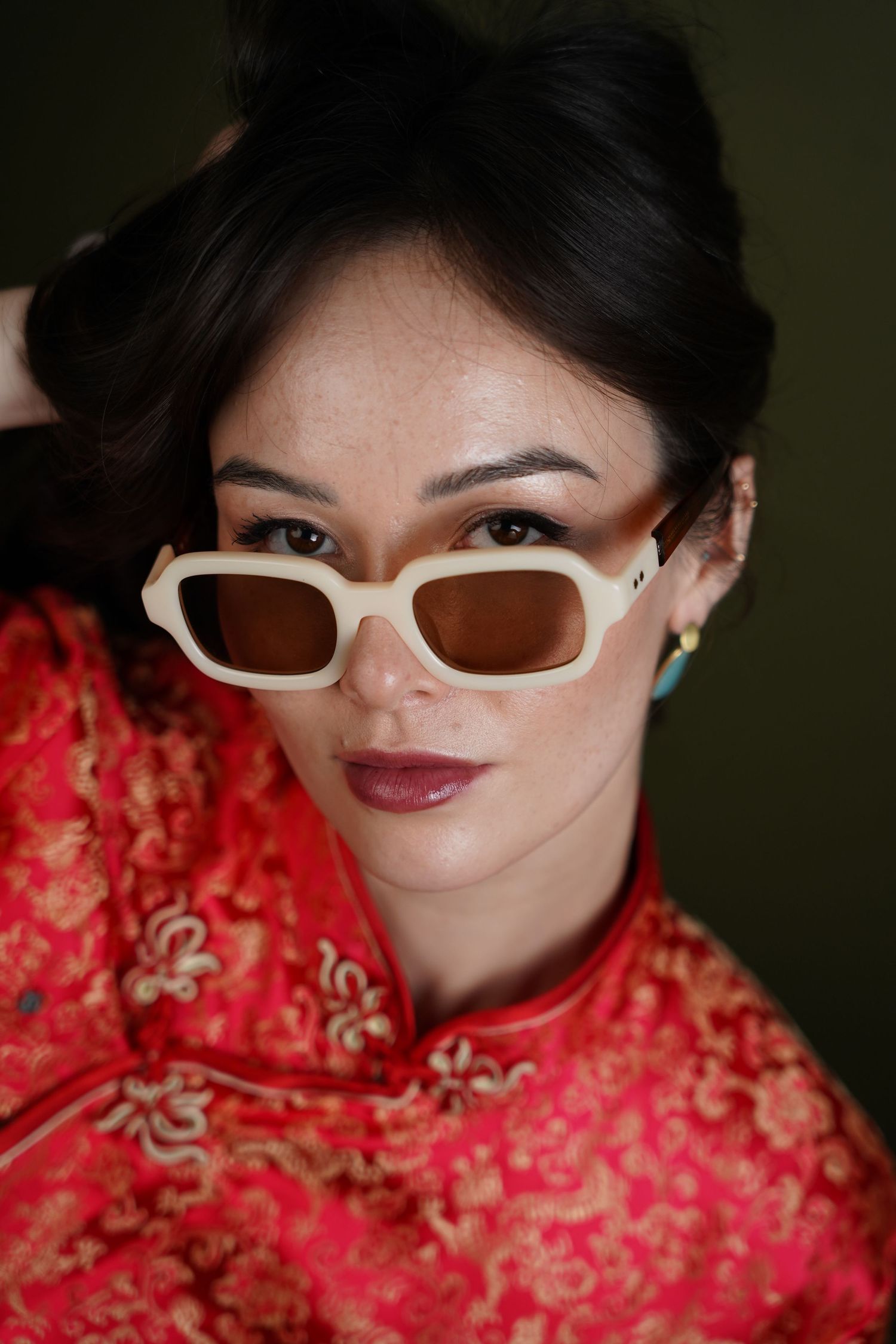 Woman in red floral cheongsam wearing cream rectangular sunglasses, posing confidently against dark background.