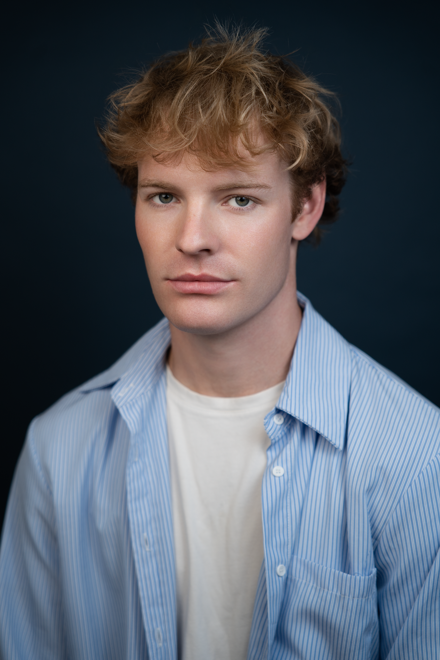 Young man with curly red hair wearing a blue striped shirt over white tee, posing against dark background.