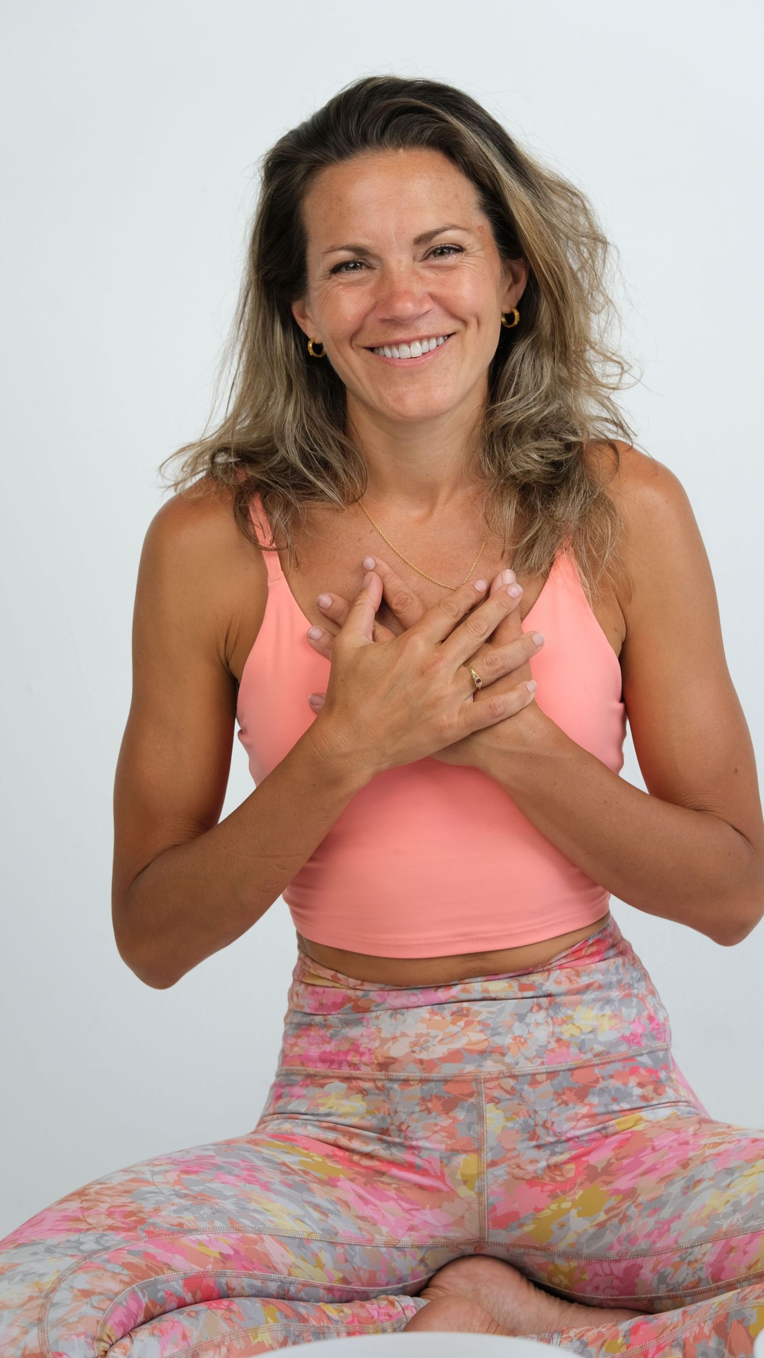 Smiling person in coral sports bra and floral yoga pants sits cross-legged with hands clasped to chest in meditation pose.