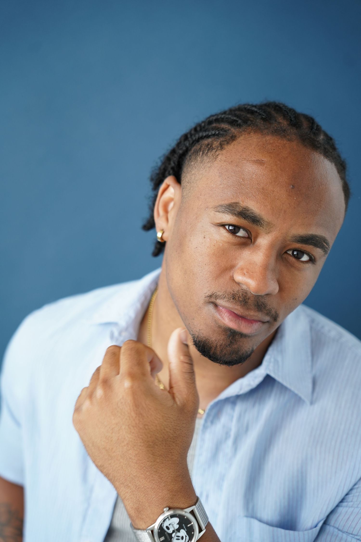 A young Black man with braids wearing a light blue shirt and watch poses confidently against a blue background.