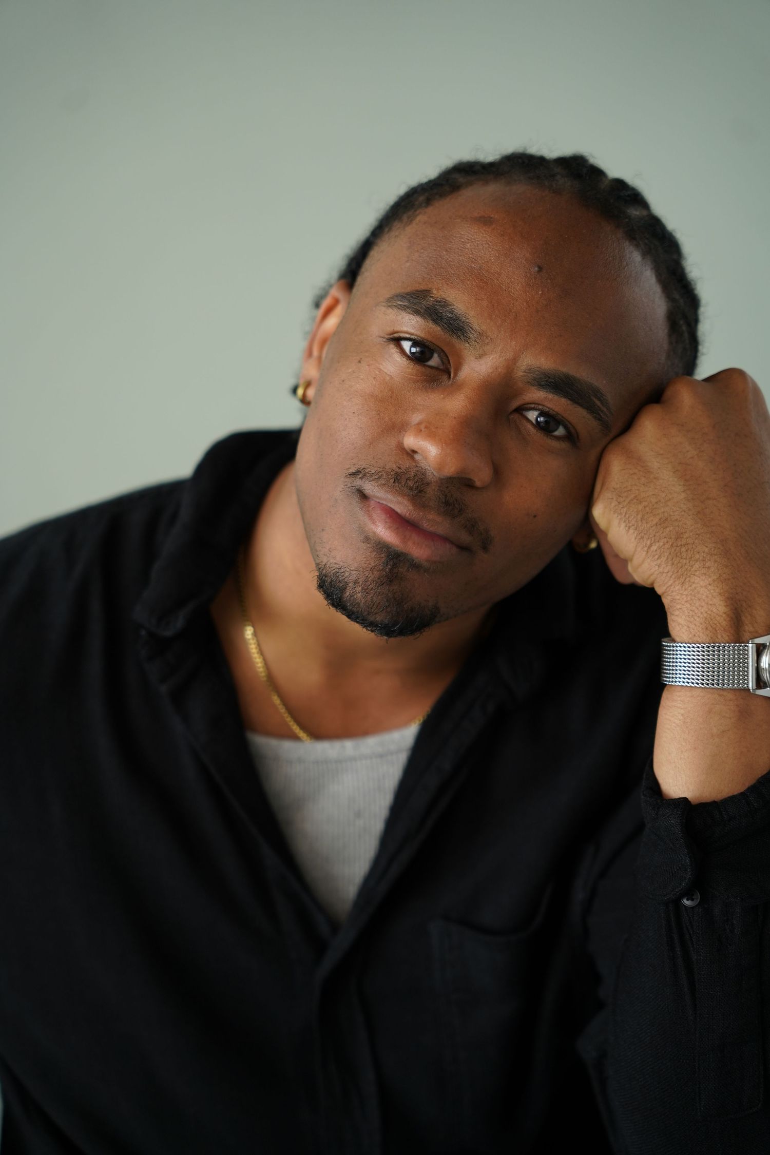 Young Black man in black shirt resting head on hand, wearing gold necklace and silver watch, thoughtful expression.