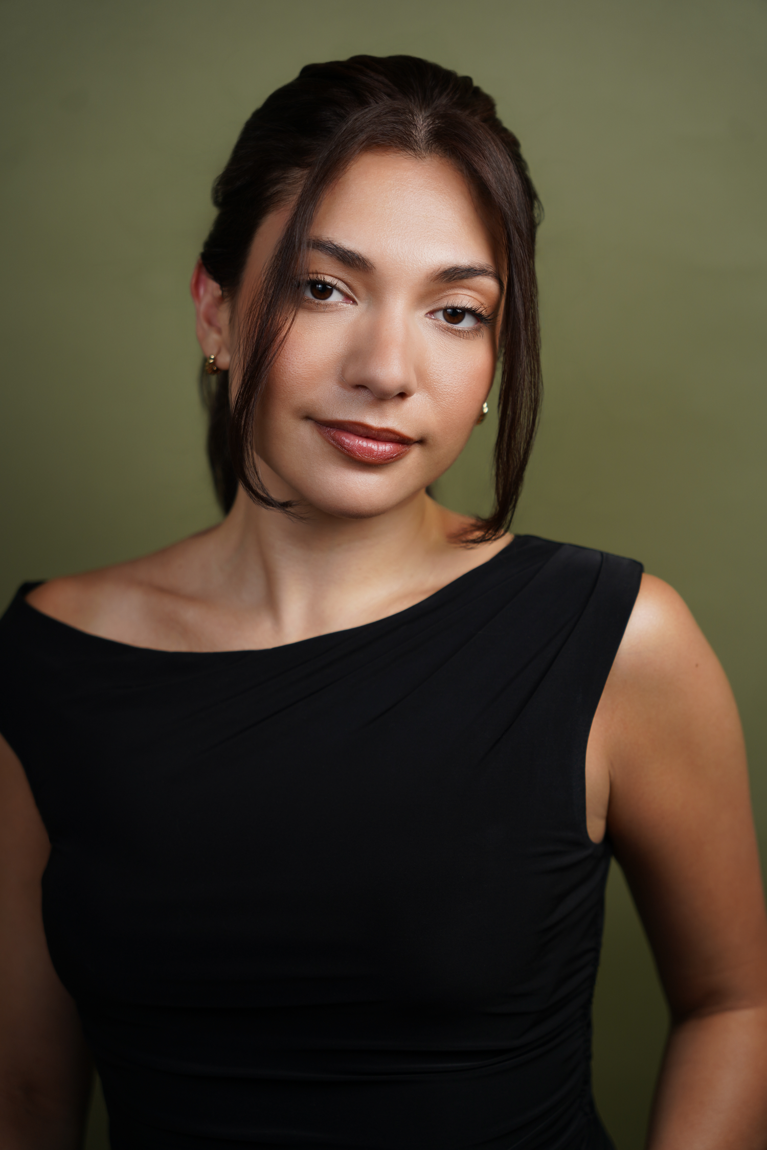 Young woman with dark hair in black off-shoulder dress poses against olive green background in professional portrait.