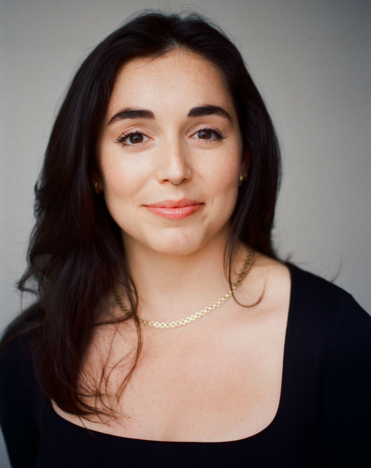 Professional headshot of a person with long dark hair wearing a black top and delicate gold necklace against a light background.
