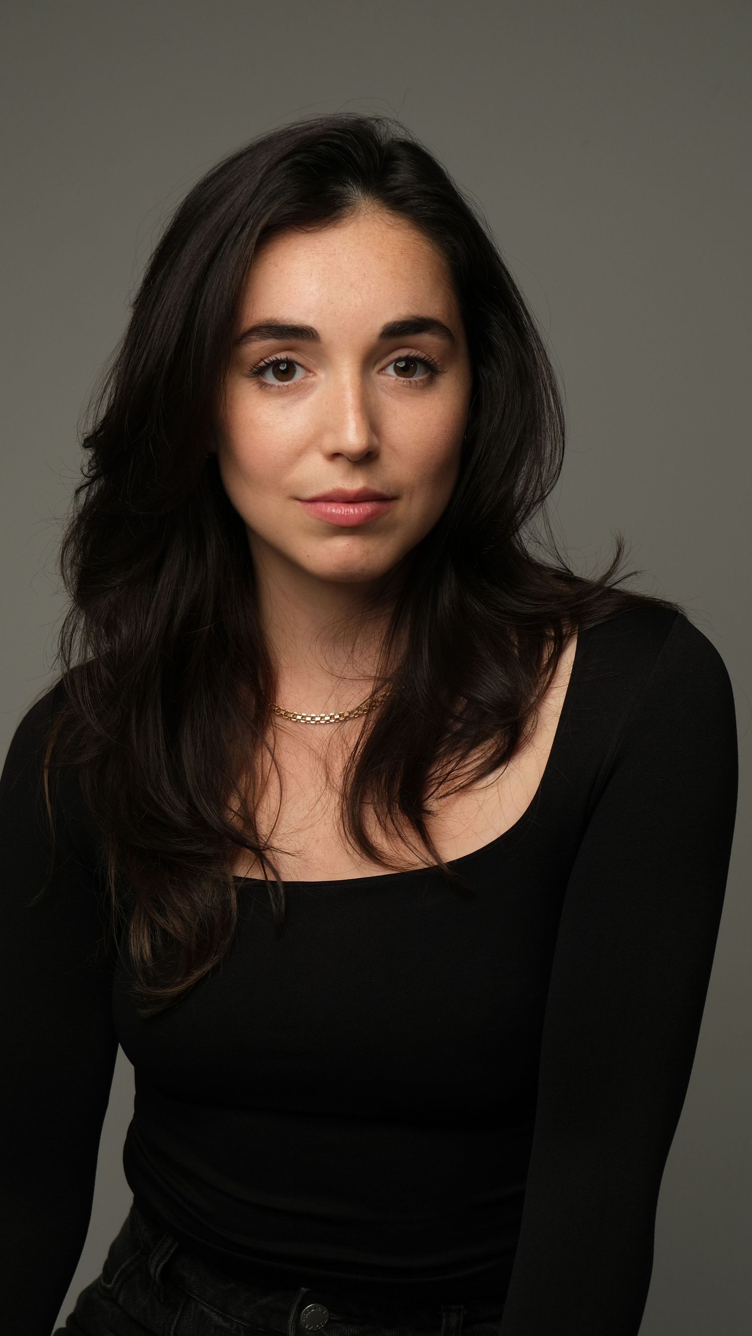 Portrait of a woman with long dark hair wearing a black top and delicate necklace against a neutral background.