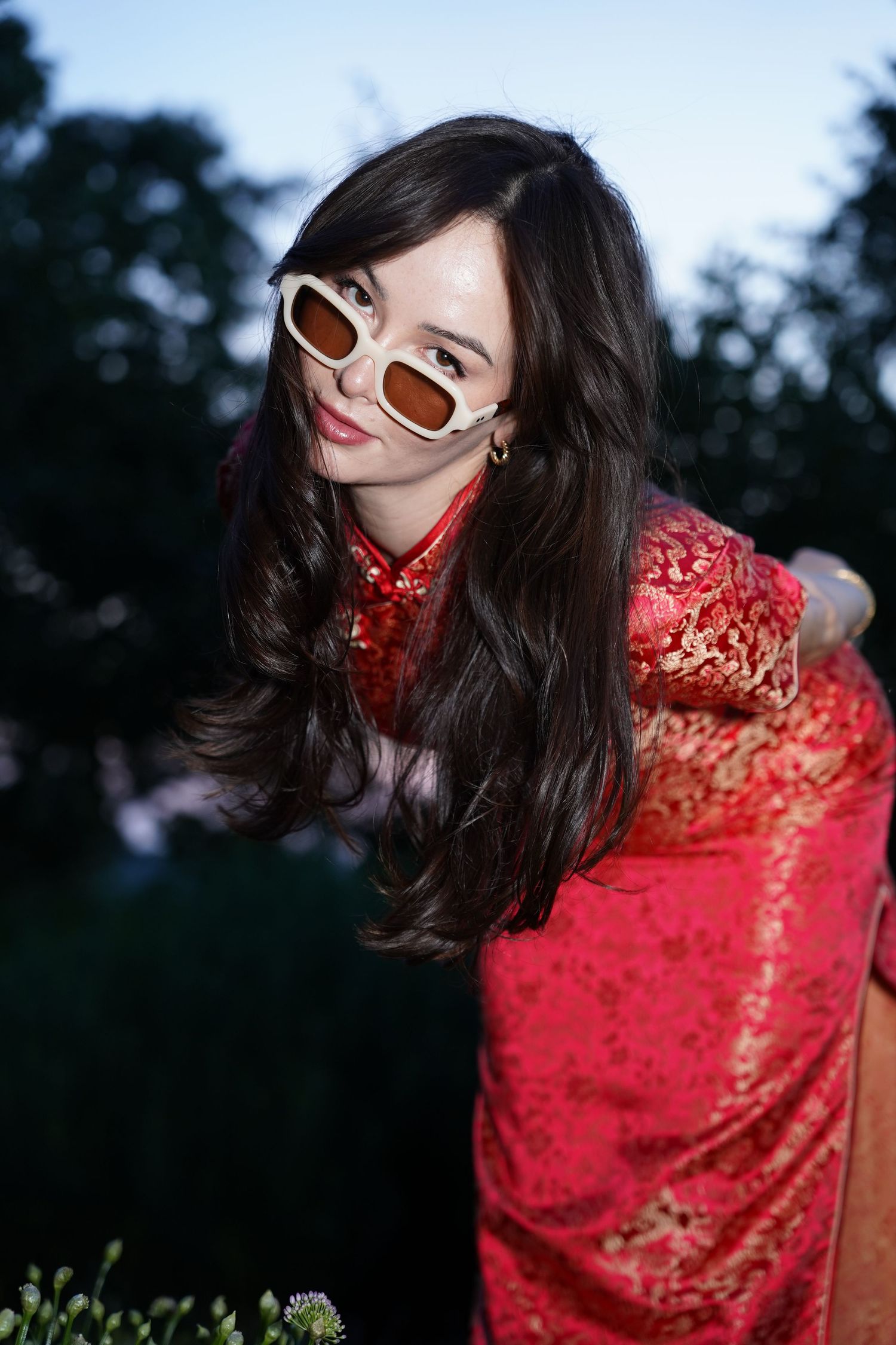 Young woman in red floral qipao and white sunglasses poses outdoors at dusk with dark trees behind her.