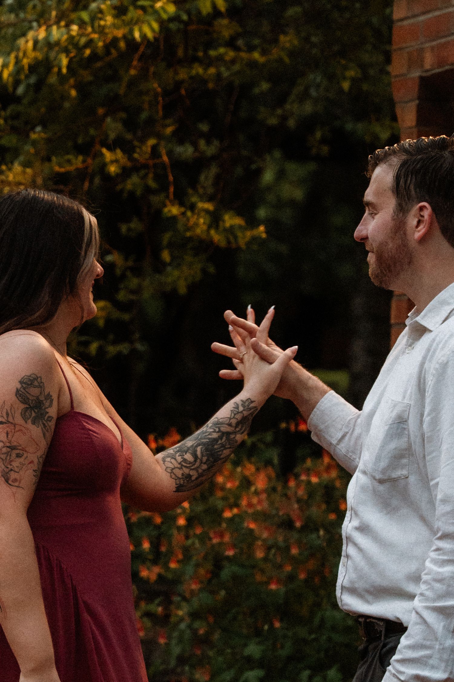 Couple admiring engagement ring outdoors, woman in burgundy dress with tattoos, man in white shirt, autumn foliage background.
