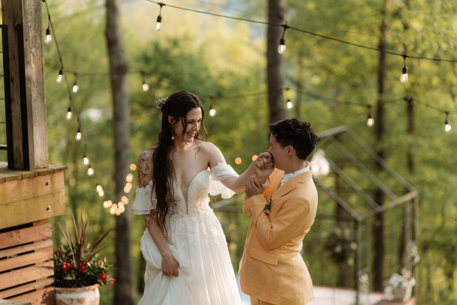 Groom in yellow suit kisses bride's hand during outdoor wedding ceremony with string lights and forest backdrop.