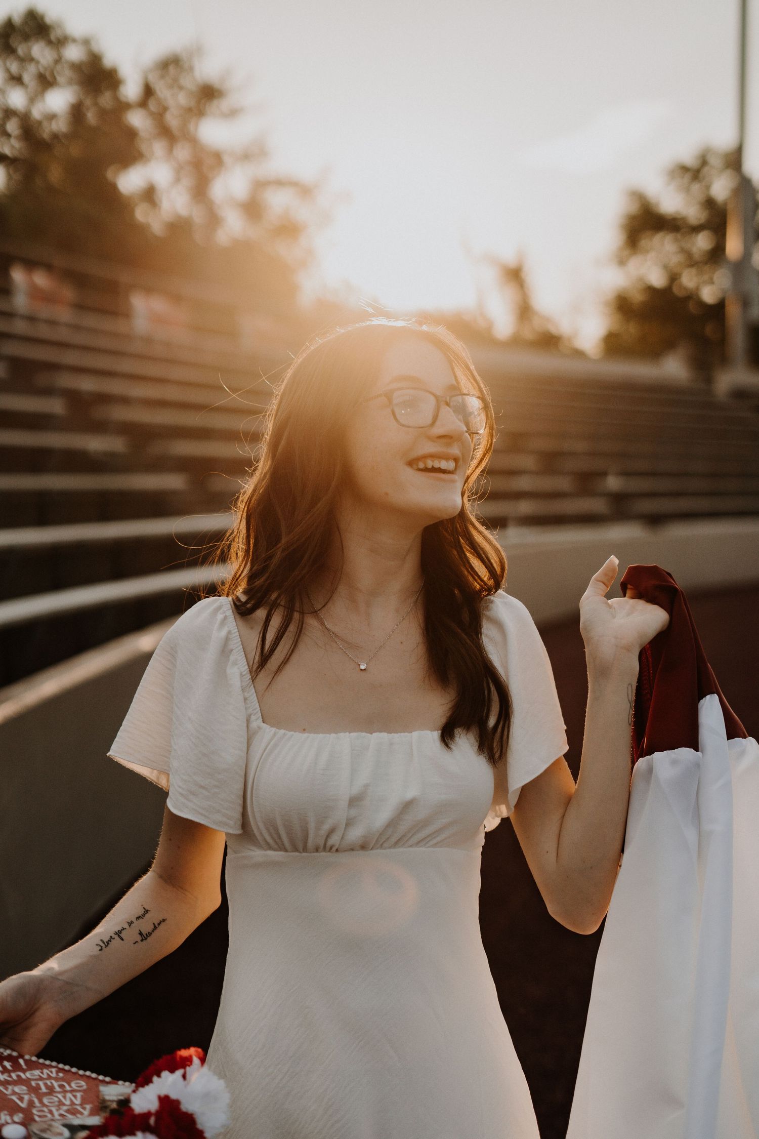 High school graduate in white dress laughing during golden hour sunset on outdoor bleachers.