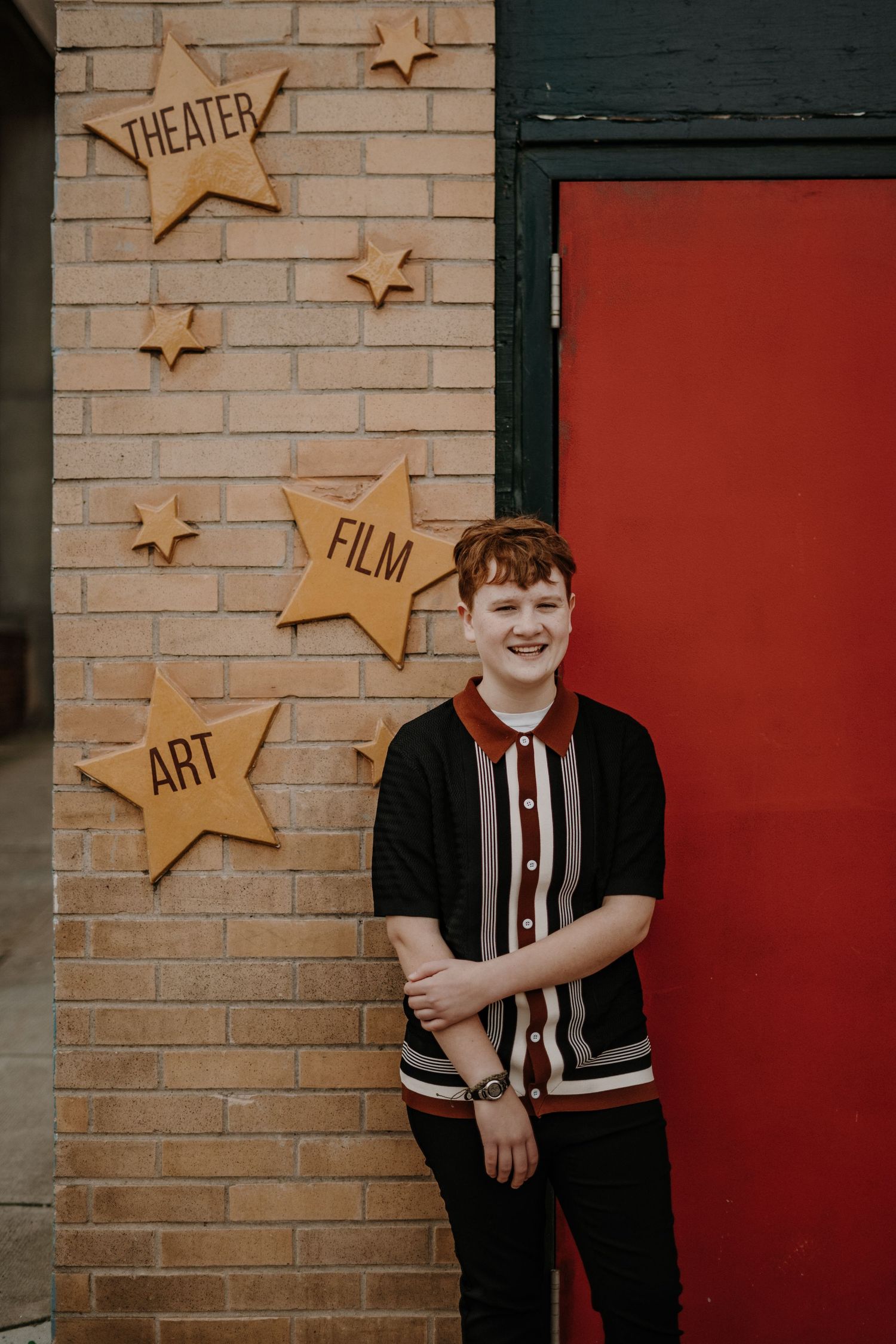 Senior portrait of a person standing by a brick wall decorated with wooden stars labeled Theater, Film, and Art.