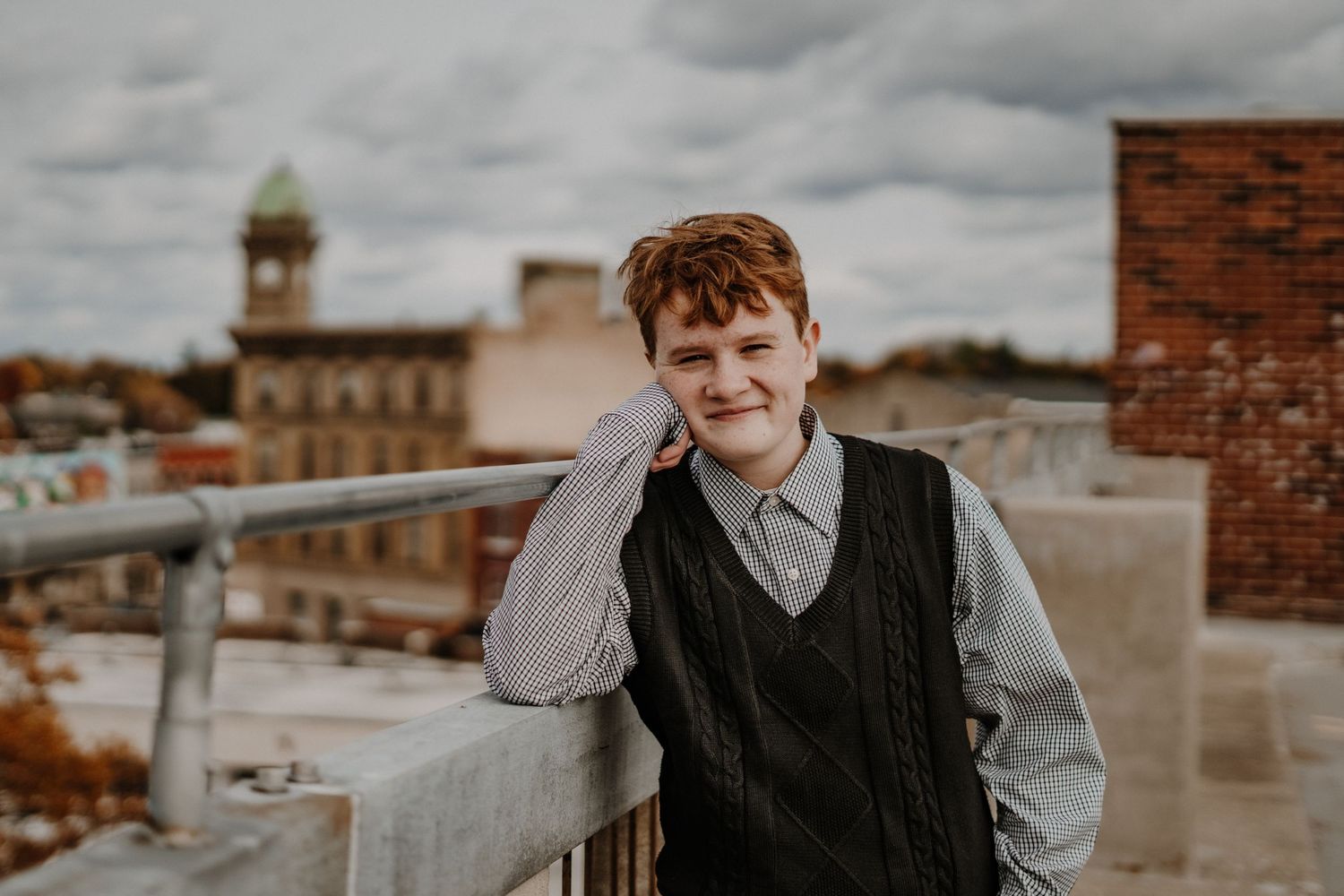 Senior portrait of person in vest sitting on urban rooftop with cityscape and cloudy sky in background.