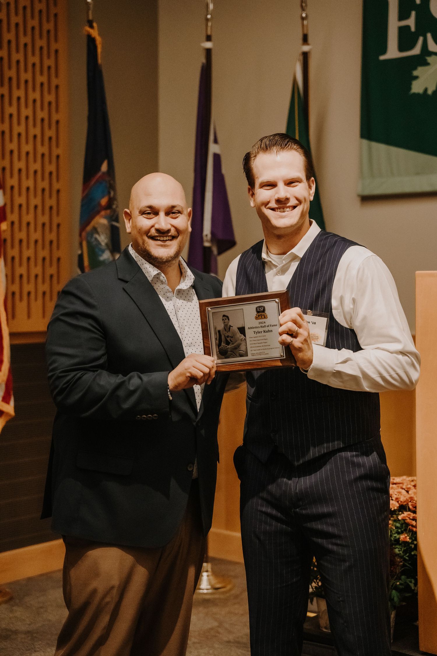 Two people in formal attire displaying an award plaque during a ceremony presentation.