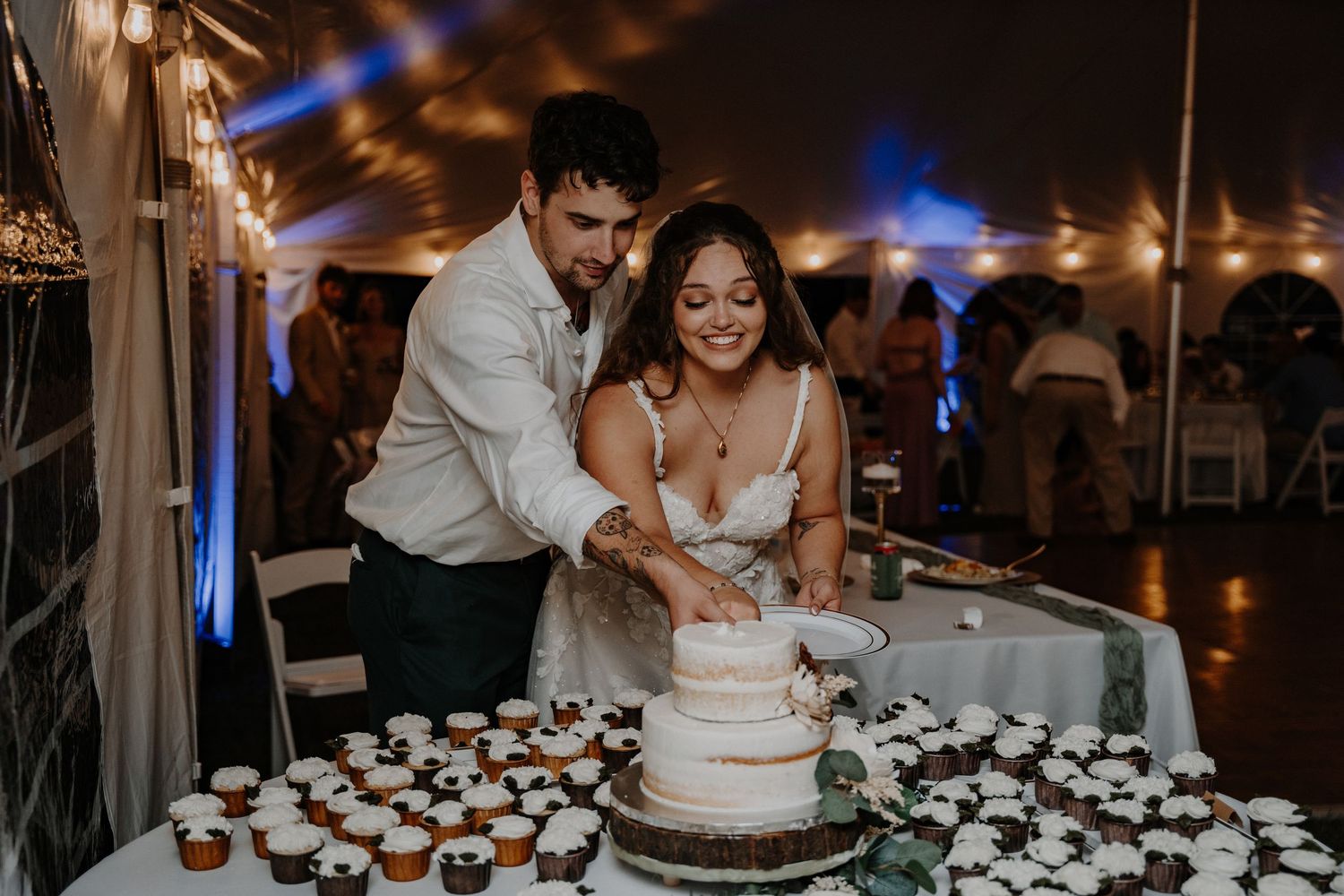 Newlyweds cutting their wedding cake together under string lights at evening reception.