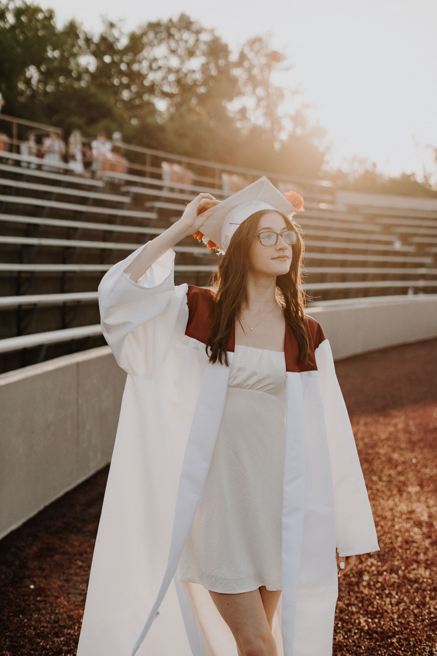 High school Graduate in white dress and cap posing on stadium bleachers during sunset.