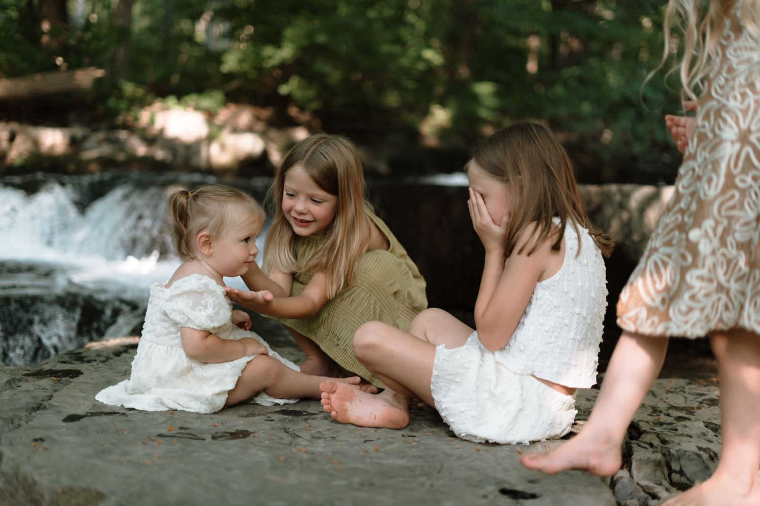 Three young girls sitting on rocks near a waterfall, laughing and playing together outdoors.