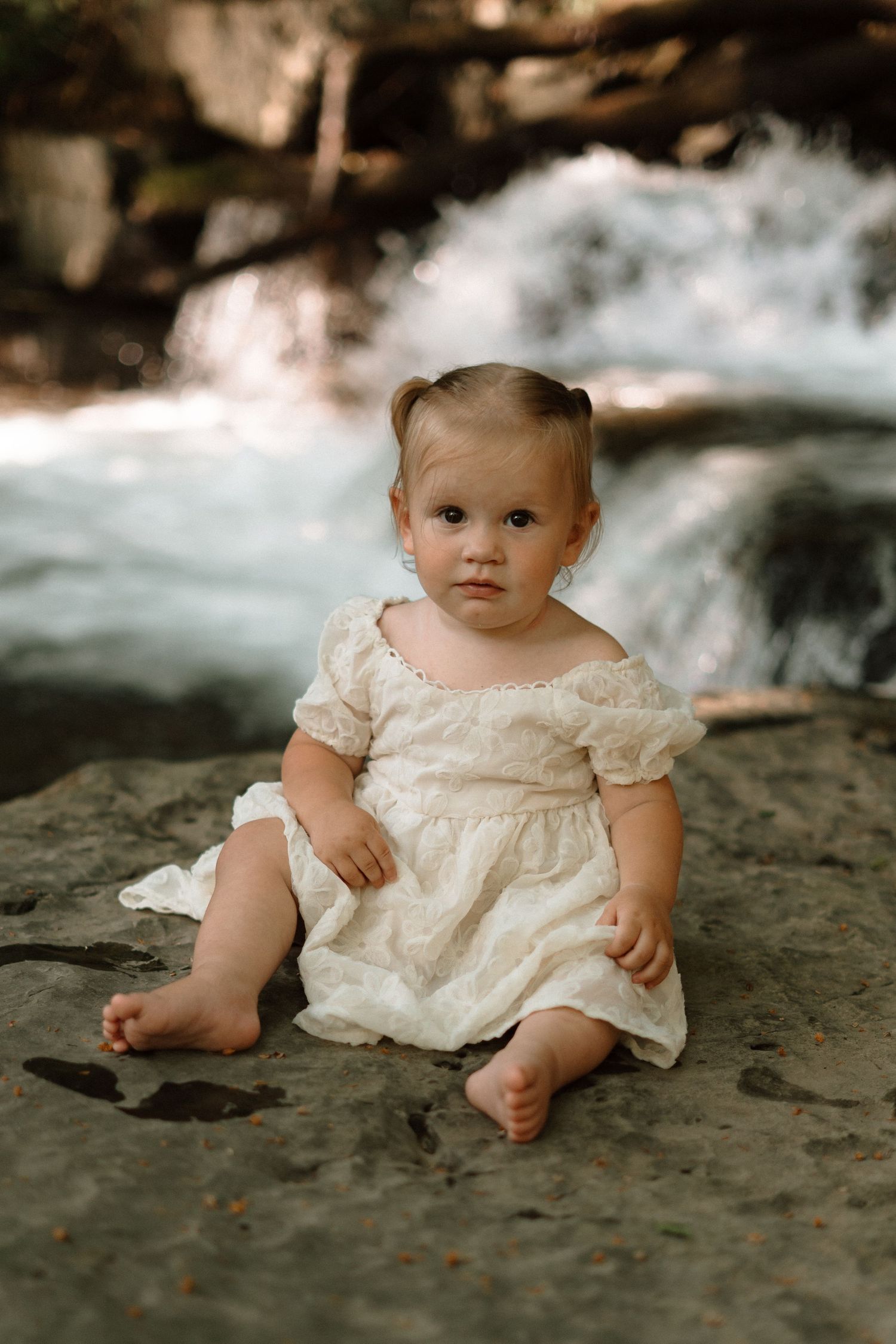 A toddler girl in a white lace dress sits barefoot on a rock near a flowing waterfall in nature.