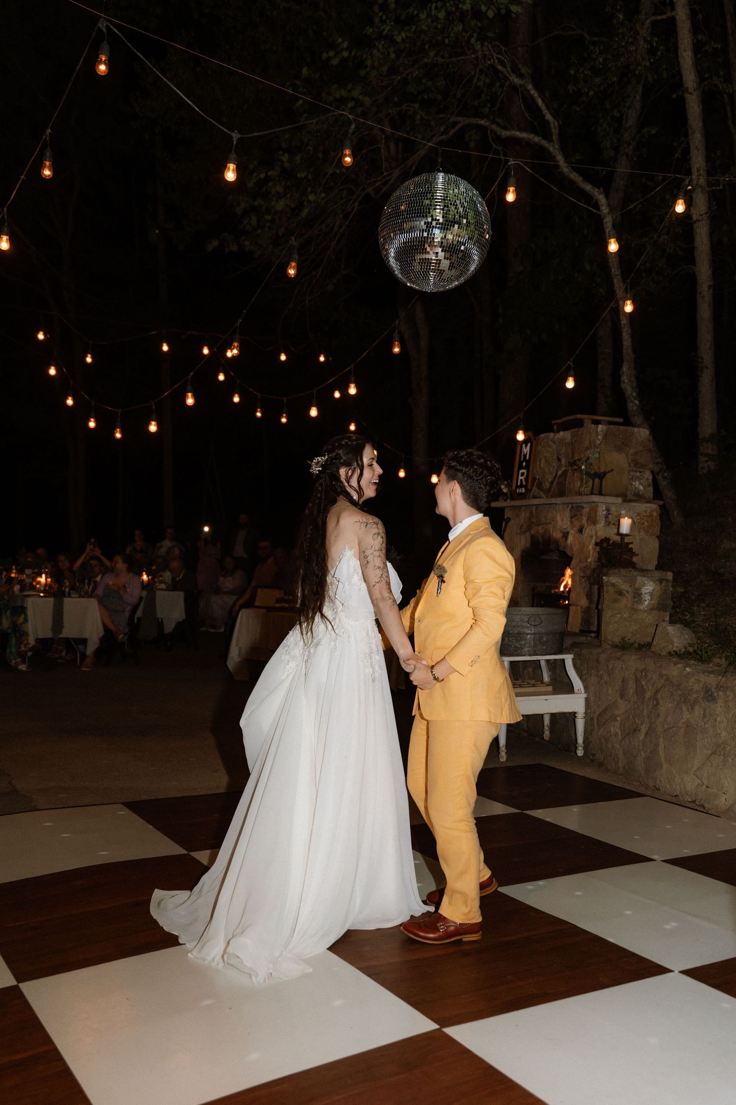 Bride and groom share first dance on checkered floor under disco ball and string lights at outdoor wedding reception.