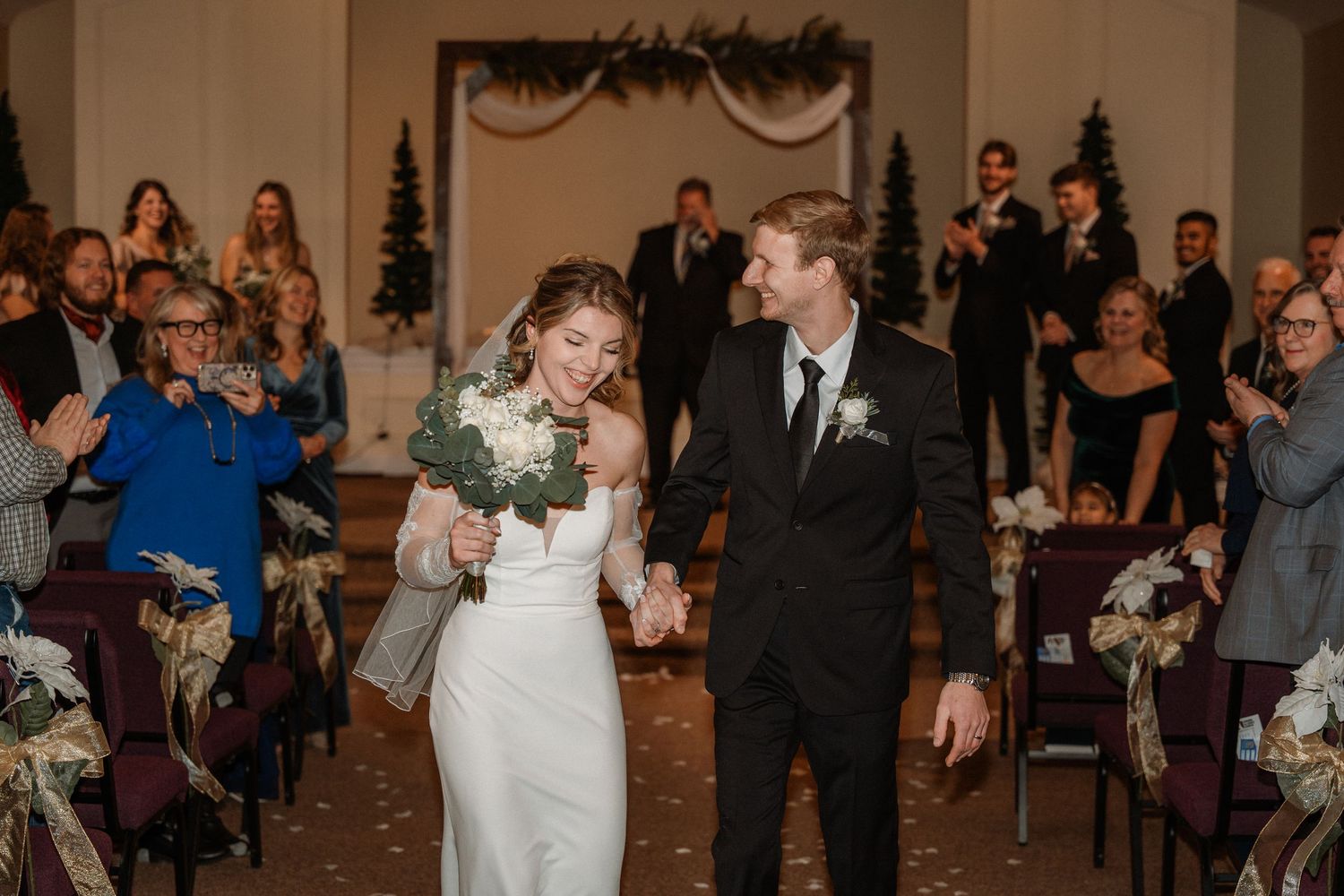 Newlywed couple walks down the aisle hand in hand, bride in white gown holding bouquet, guests applauding in decorated venue.