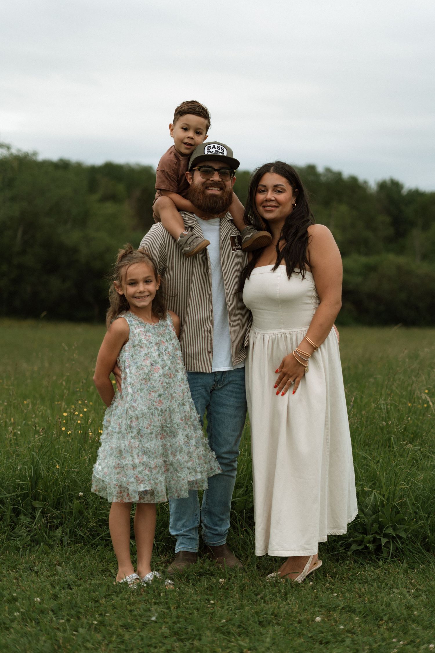 Happy family of four posing outdoors in a green field, parents with young daughter and toddler son on dad's shoulders.