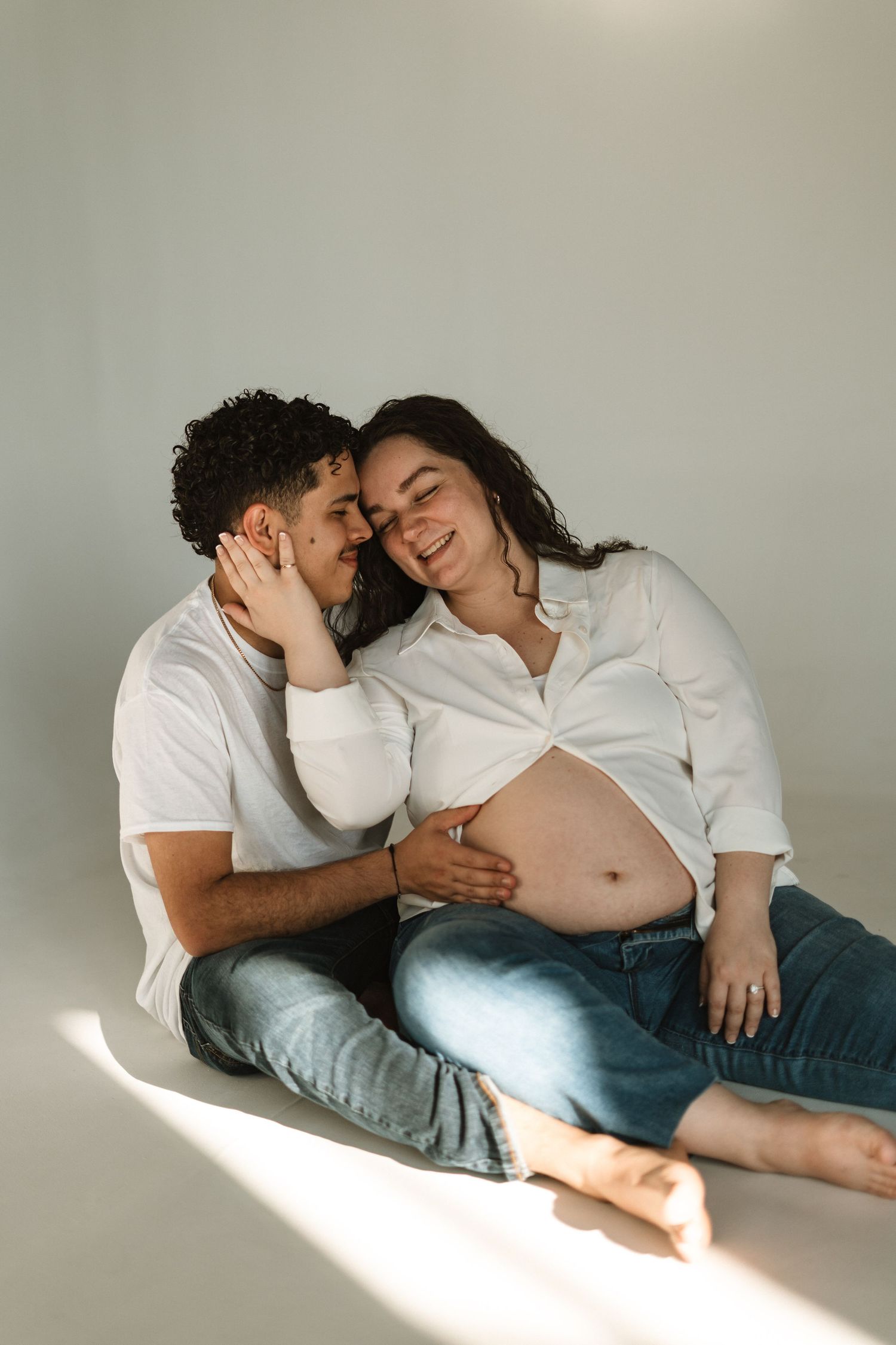 Intimate maternity photo series showing a couple in white shirts and jeans laying together in soft natural light.