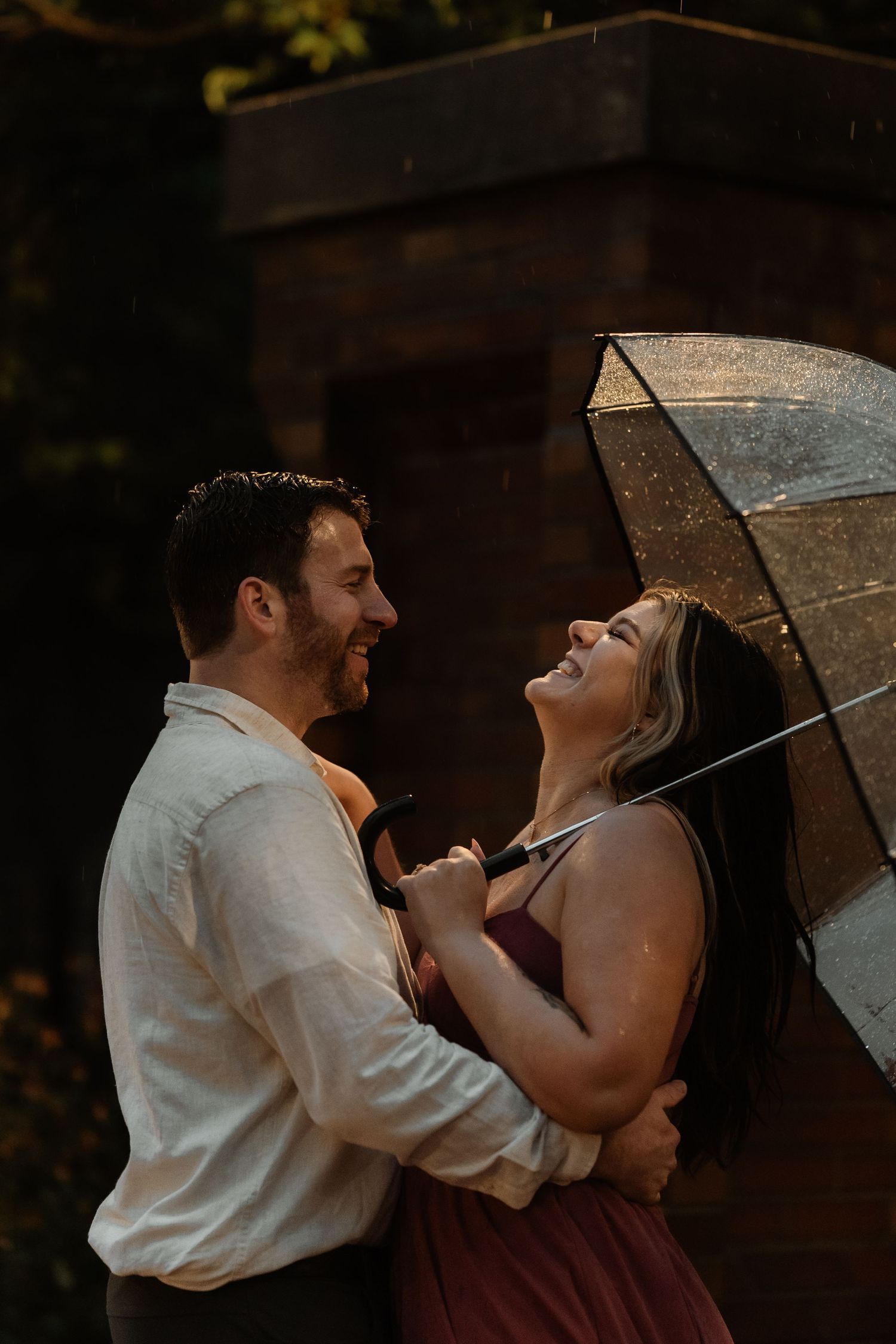 Couple laughing together in the rain under a clear umbrella during an outdoor engagement photo session.