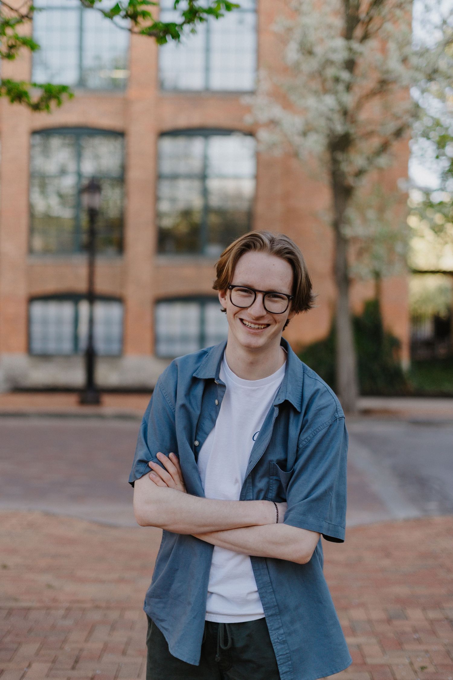 Senior portrait wearing glasses and denim jacket poses with crossed arms against a brick building.