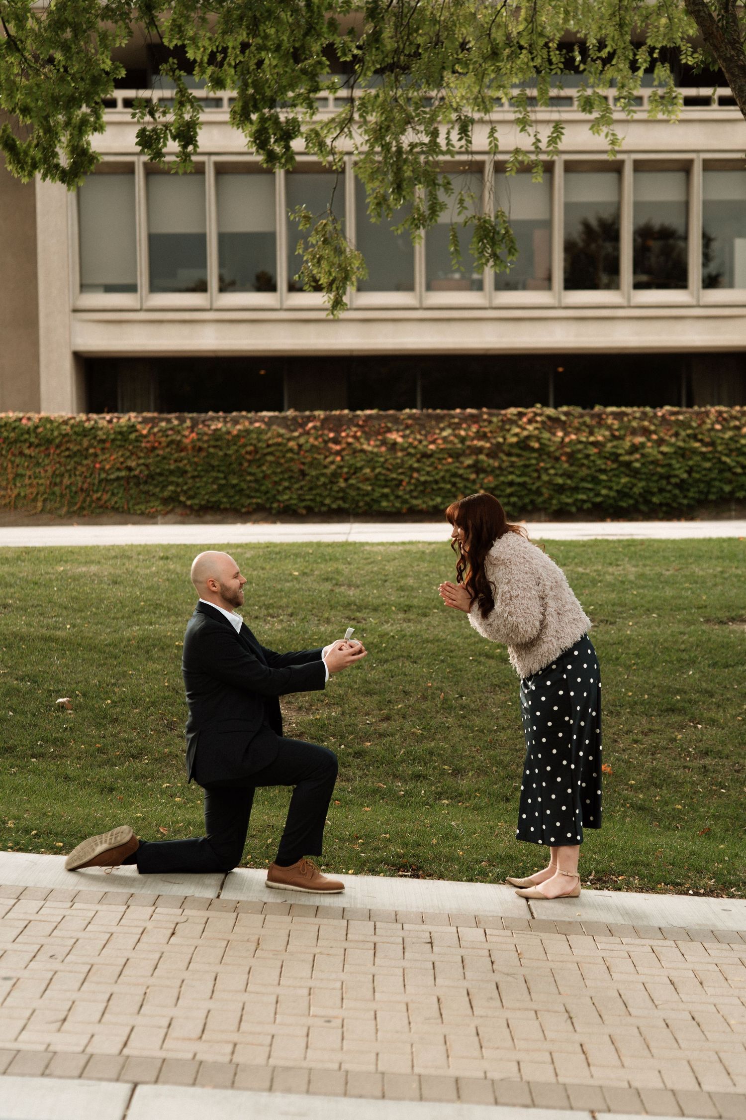 Man in black suit proposes on one knee to woman in polka dot skirt outdoors near a building.
