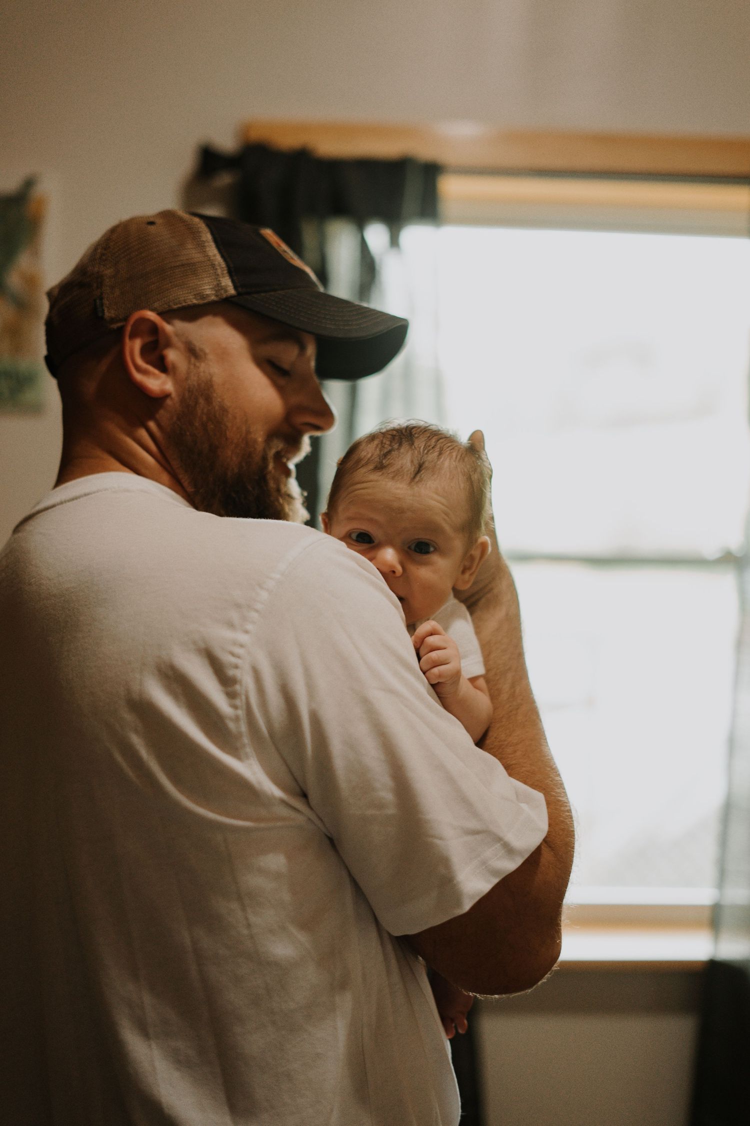 Father wearing a baseball cap holds a small infant against their chest in a warmly lit indoor setting.