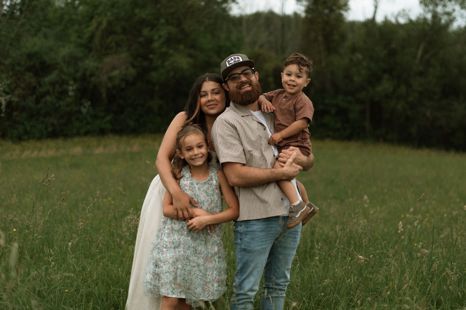 Family wearing casual outfits embraces while standing in green meadow during golden hour nature photography session.