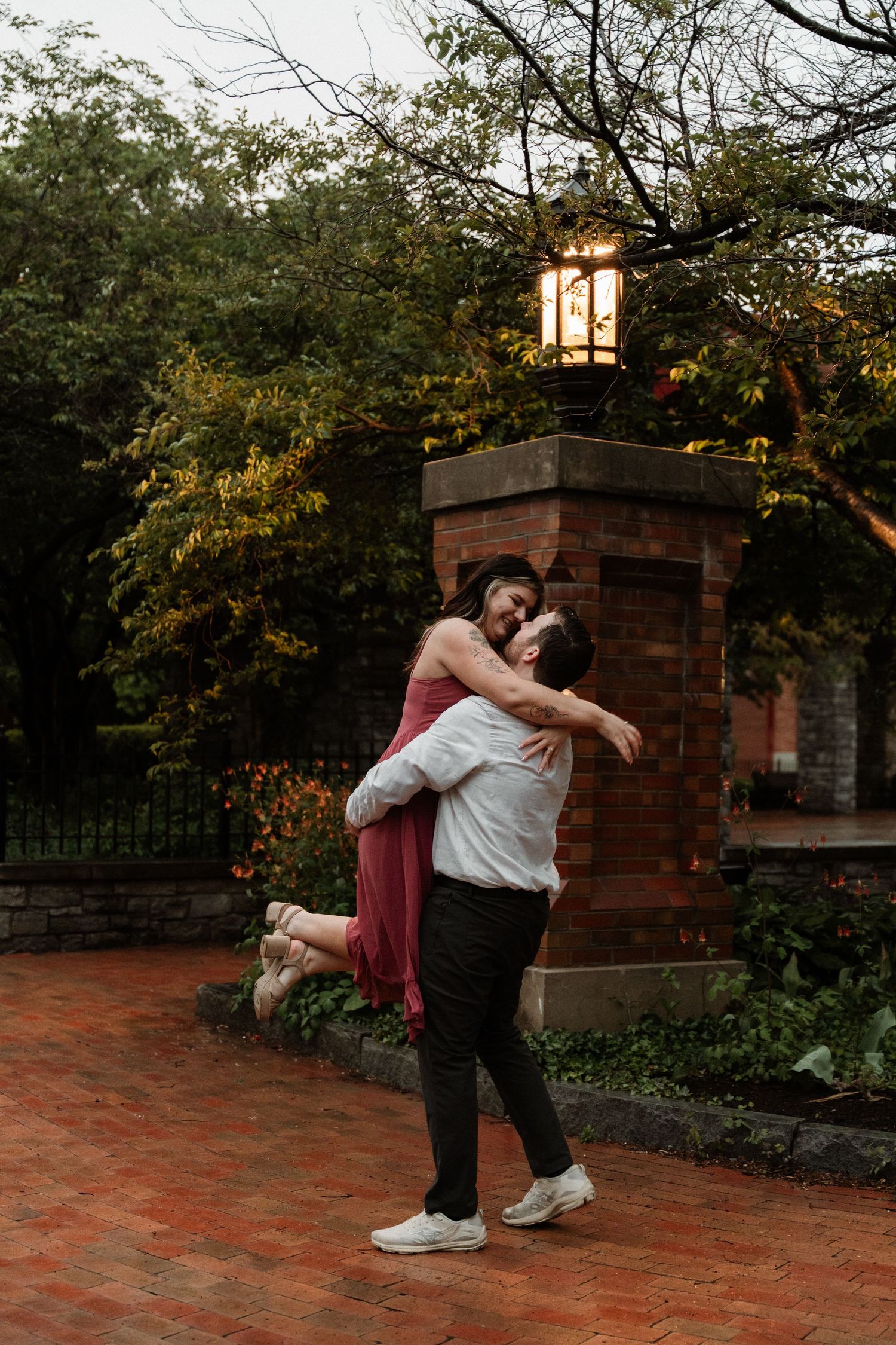 A couple shares a romantic embrace as the man lifts the woman in a red dress near a brick pillar.