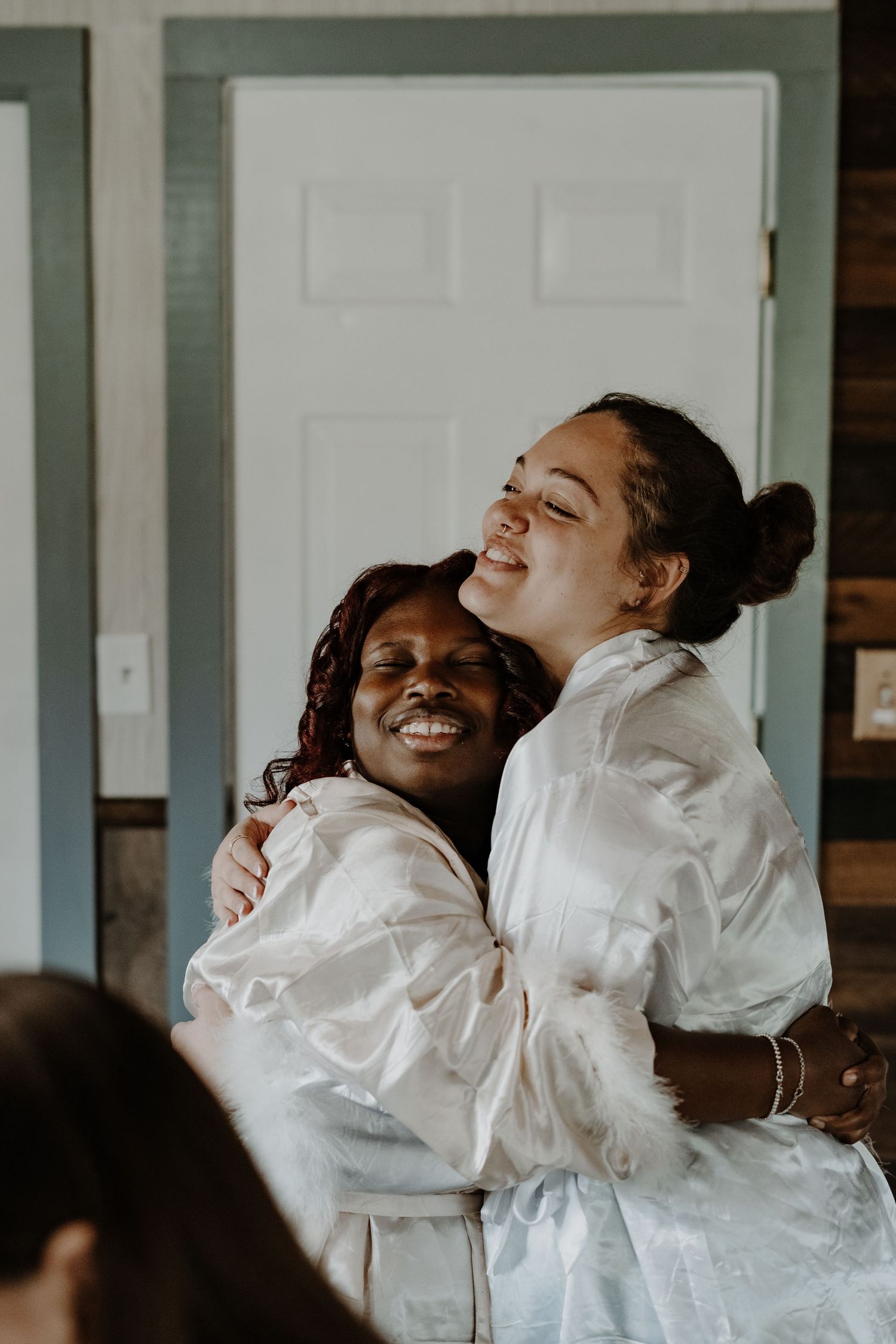 Bride and bridesmaid in white robes embracing and laughing together indoors.