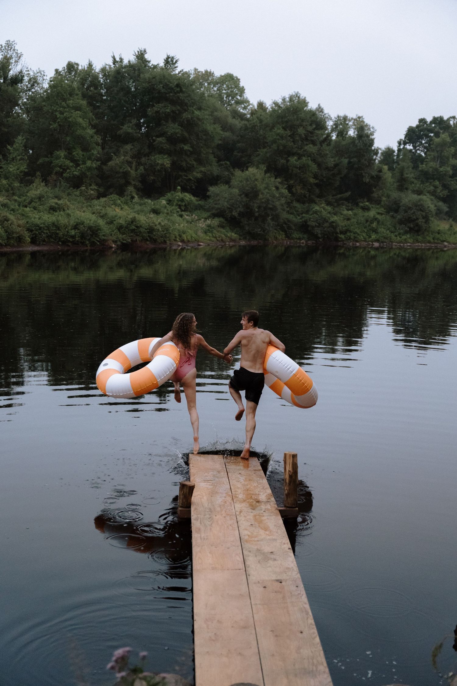 Two people attempt backflips off a wooden dock into a serene lake surrounded by pine trees on a summer day.
