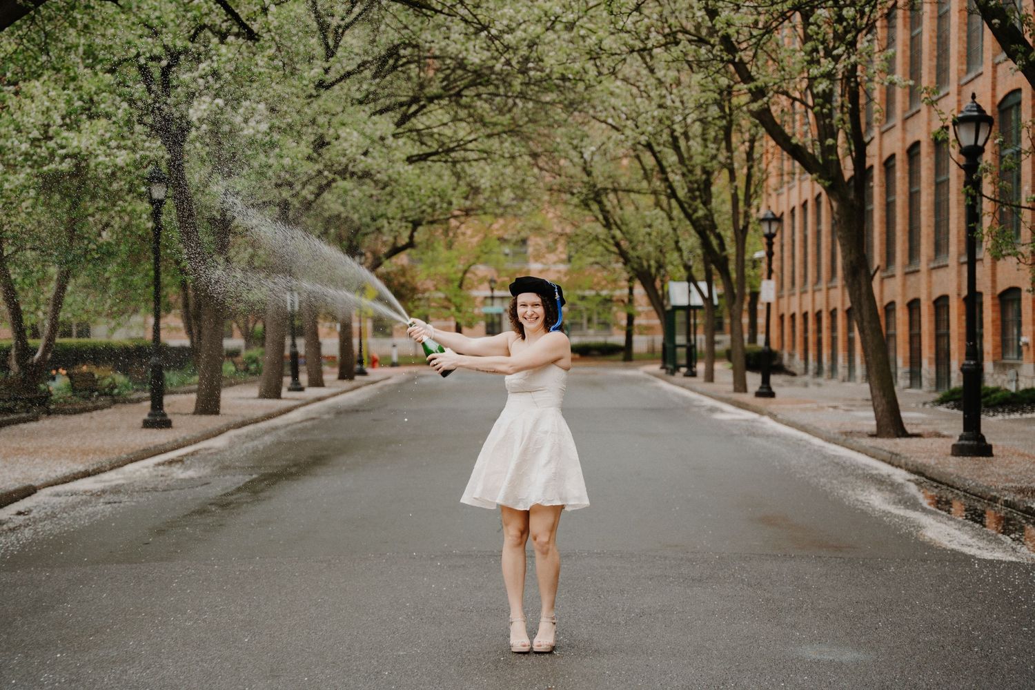 Graduation portrait of person in white dress twirling on empty city street with spraying champagne