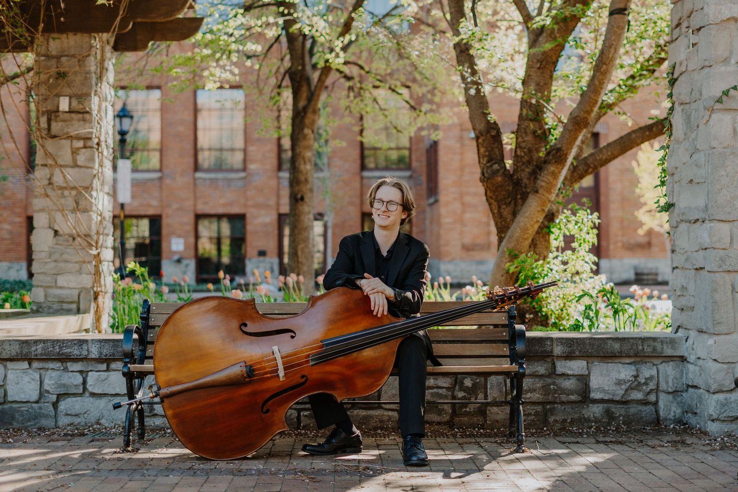 Senior portrait with upright bass in front of stone building and green trees.