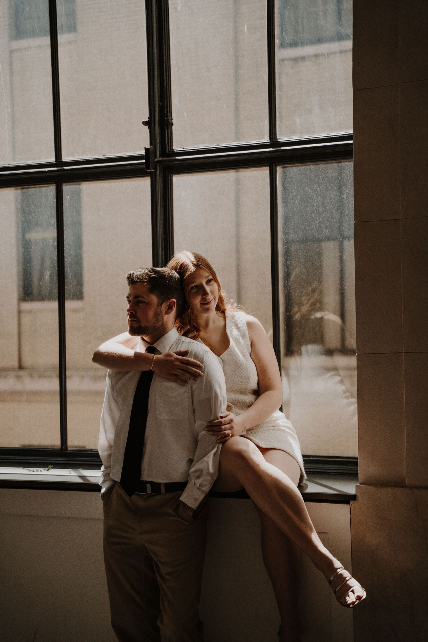 Couple poses intimately against large windows in modern industrial setting.