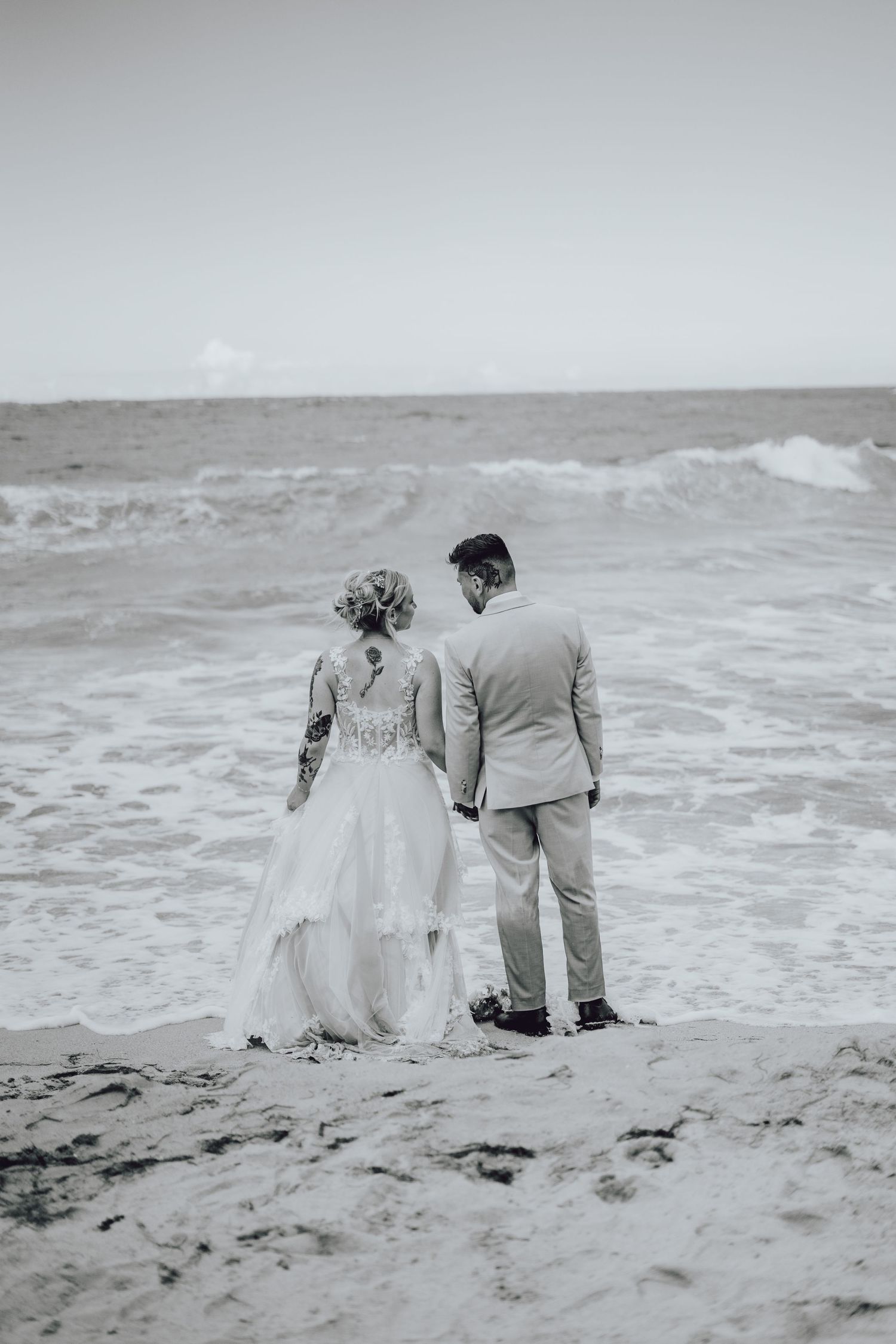 Wedding couple stands together on sandy beach with crashing waves in black and white.