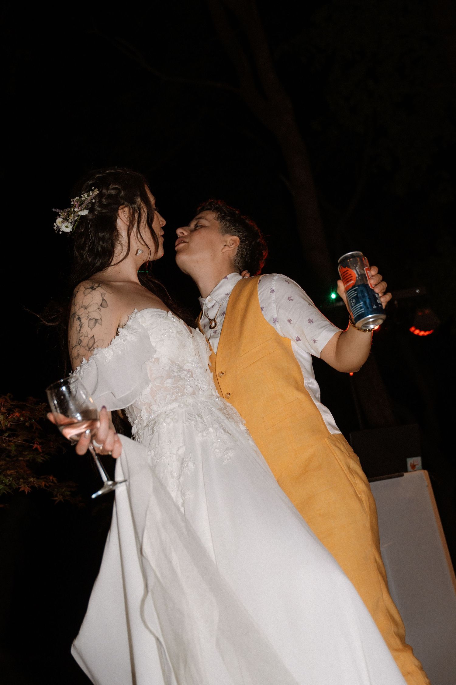 Two brides share a romantic kiss at their outdoor evening wedding reception, one in a white gown, the other in a gold vest.