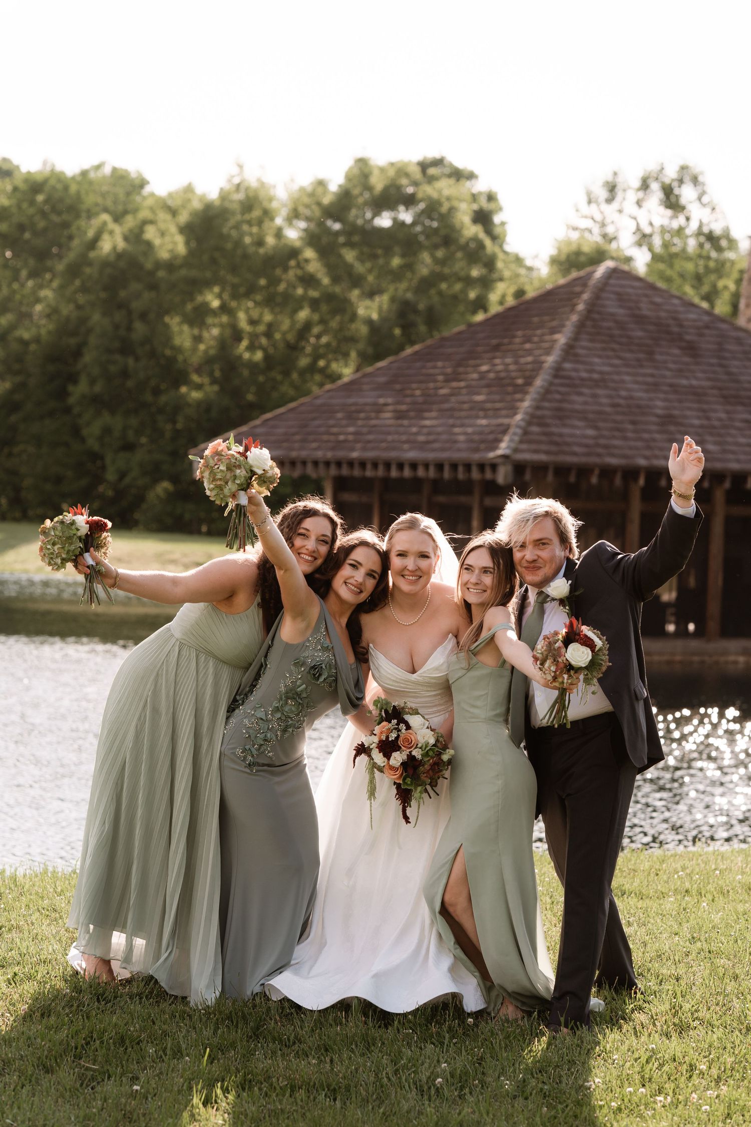 Bride in white gown poses with bridesmaids in sage green dresses and a guest by a lake at an outdoor wedding.
