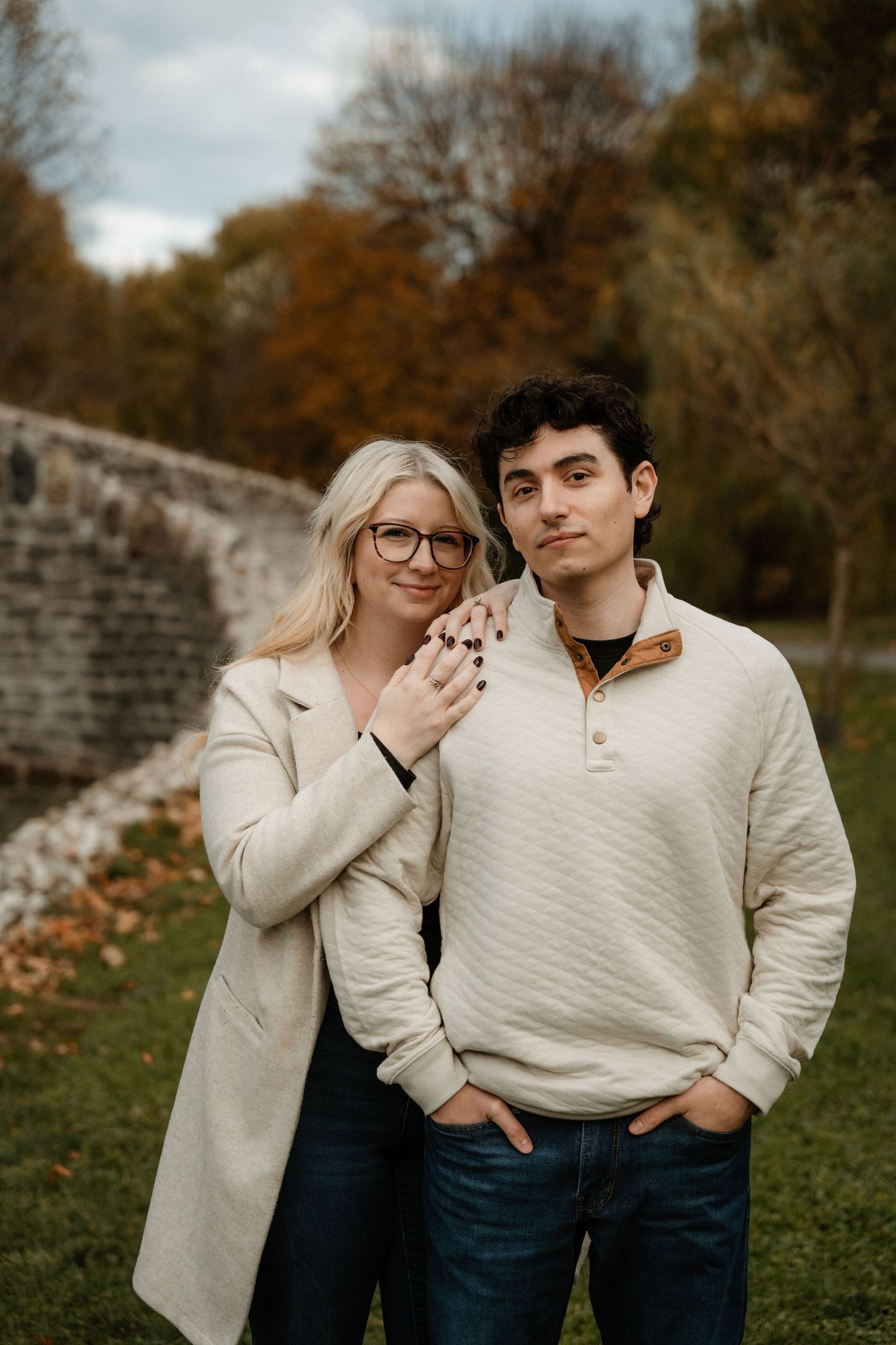 A couple poses outdoors in autumn, woman in white coat with glasses, man in cream sweater and jeans, fall foliage background.