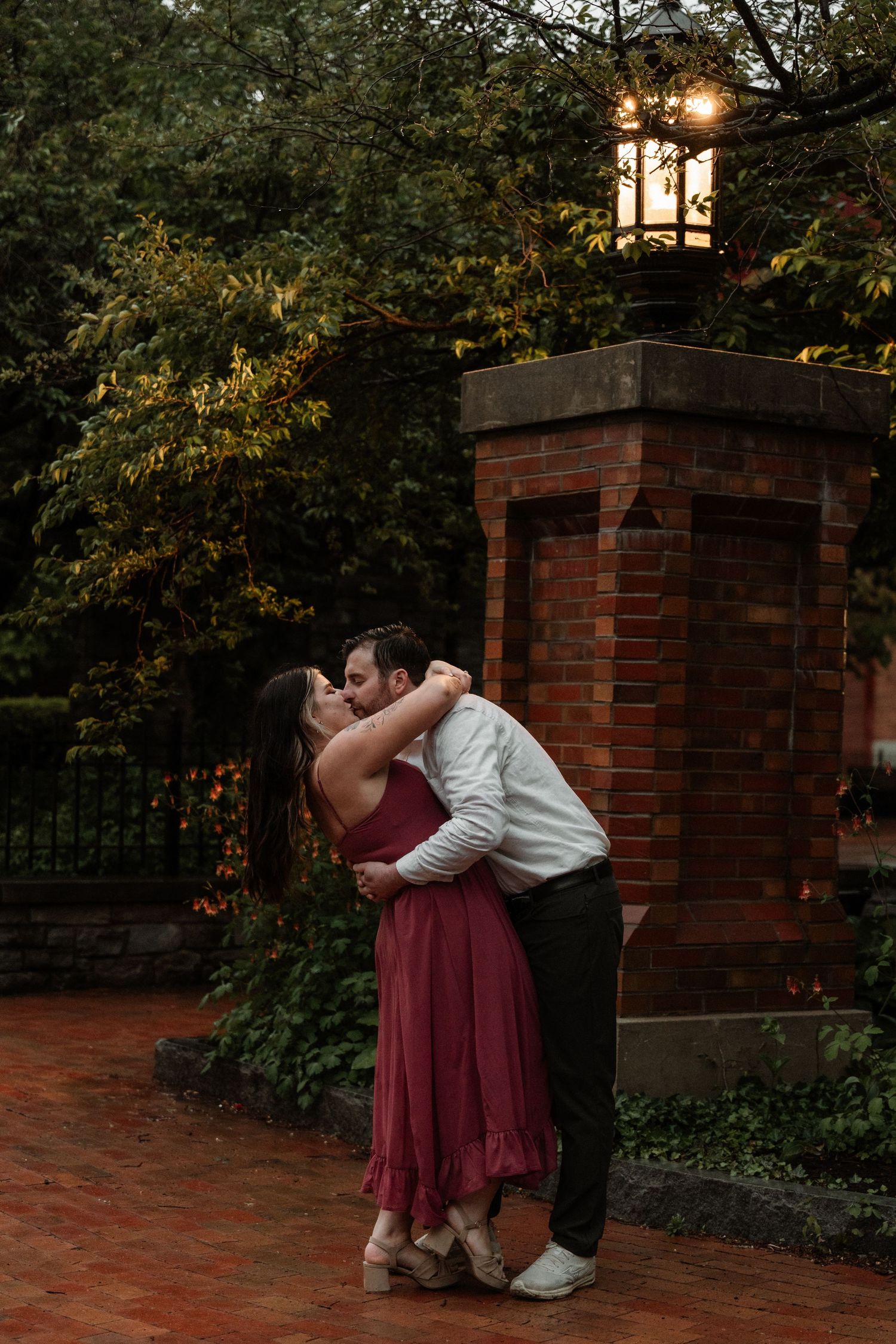 A couple shares a passionate kiss on a brick pathway near a glowing lantern surrounded by lush greenery.