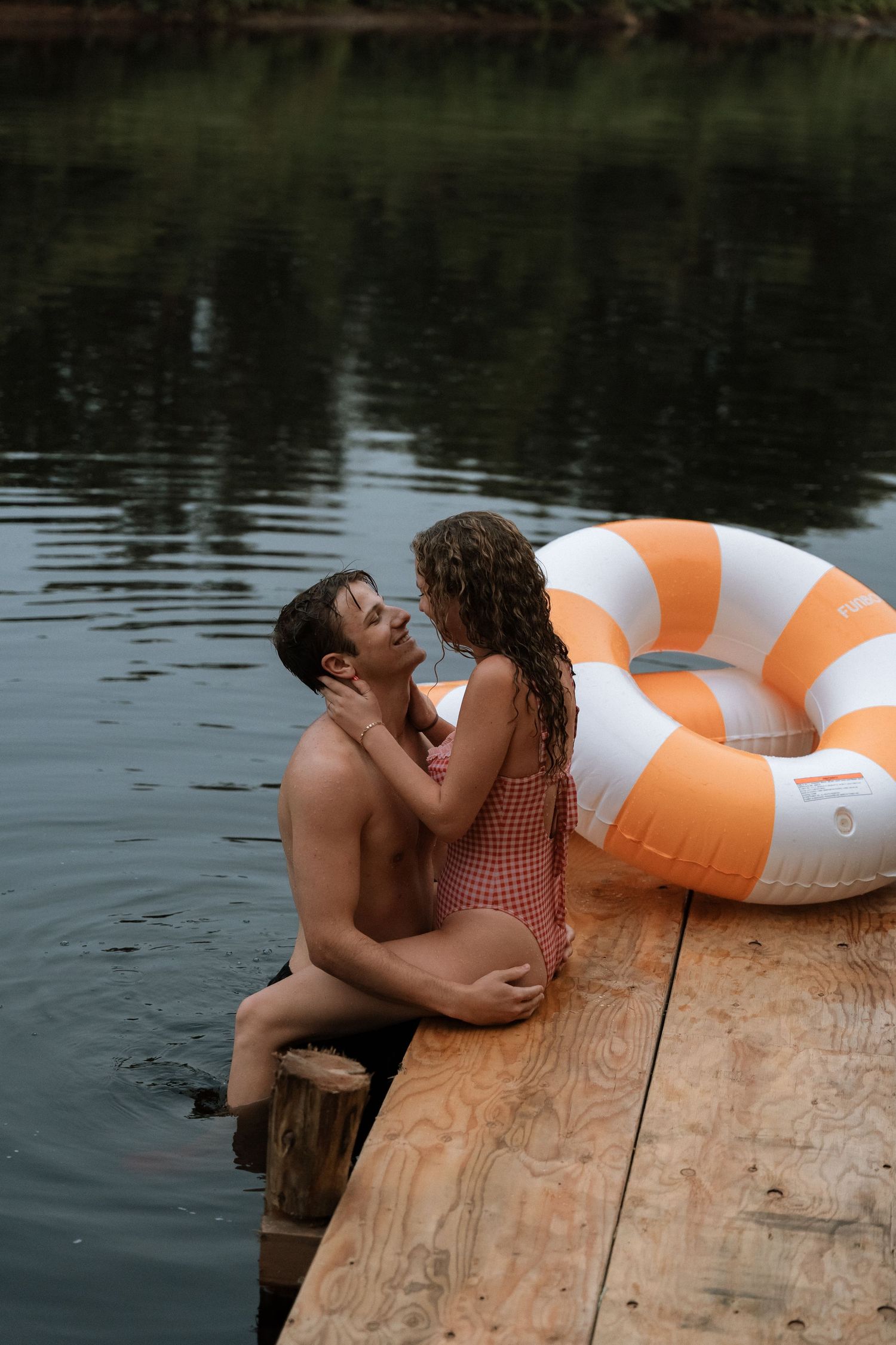 A couple shares a romantic moment on a wooden dock with an orange and white pool float beside them.