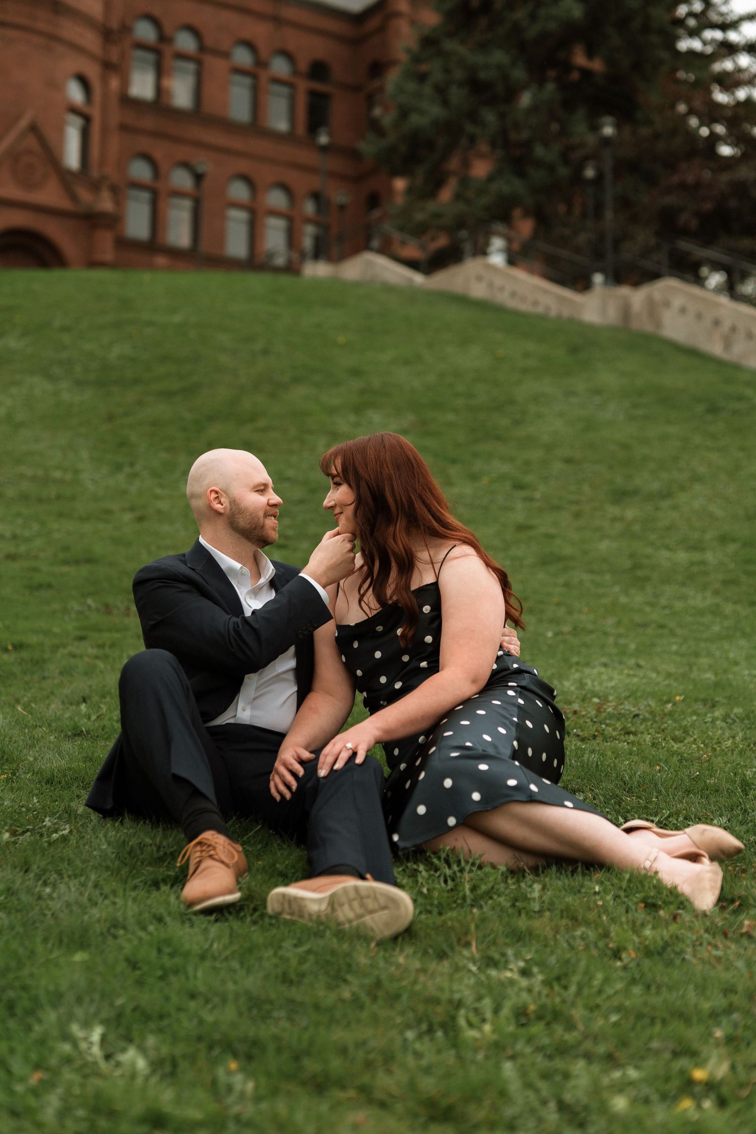 A couple shares an intimate moment while sitting on a grassy hill near a historic brick building.