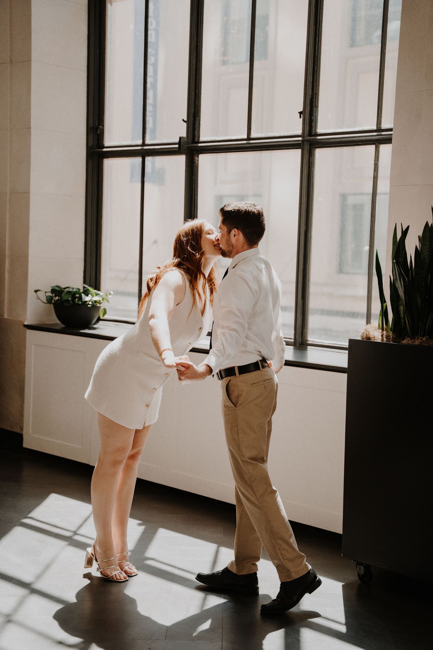 Couple sharing an intimate kiss by large industrial windows in sunlit room.