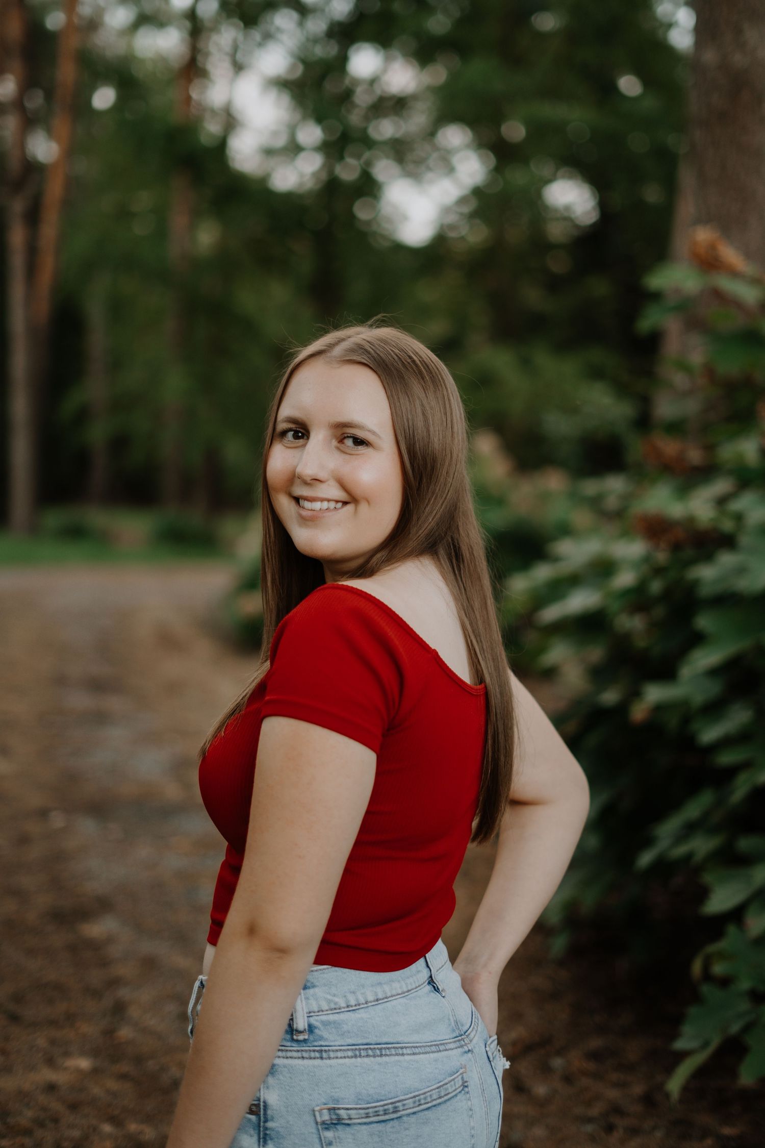 Senior portrait of person in red shirt and jeans posing against natural woodland background.