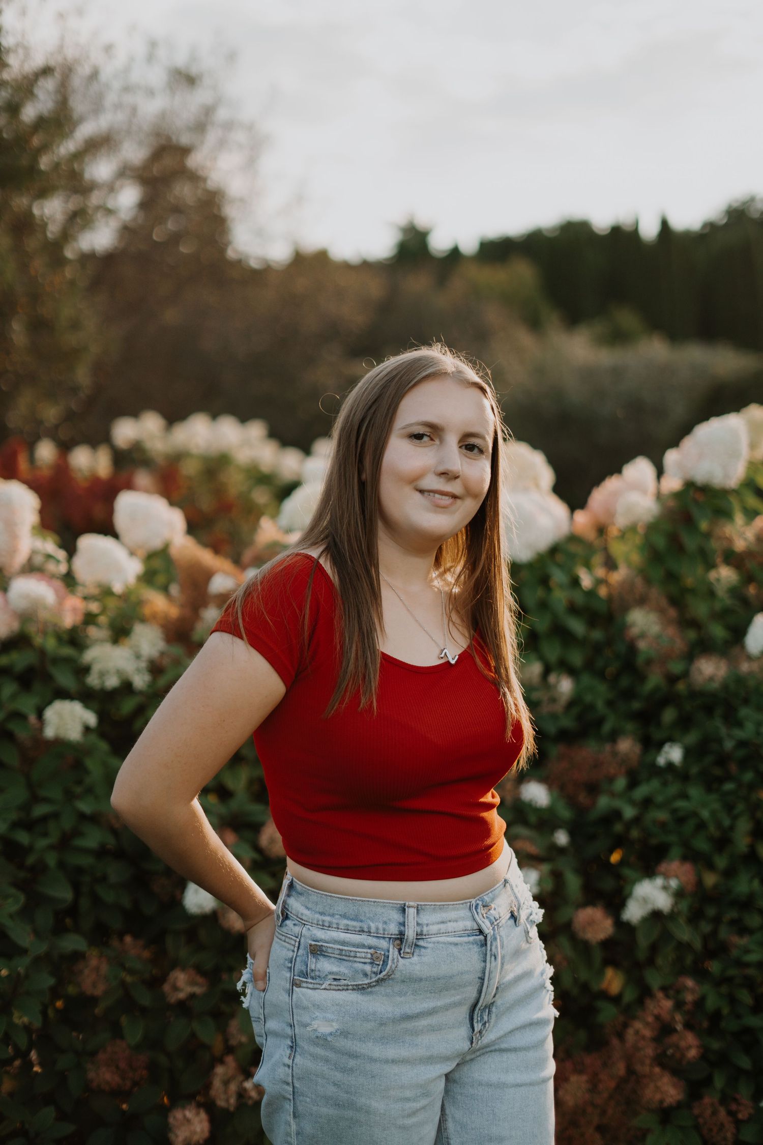 Senior portrait of person in a red crop top poses in a garden with blooming roses at sunset.
