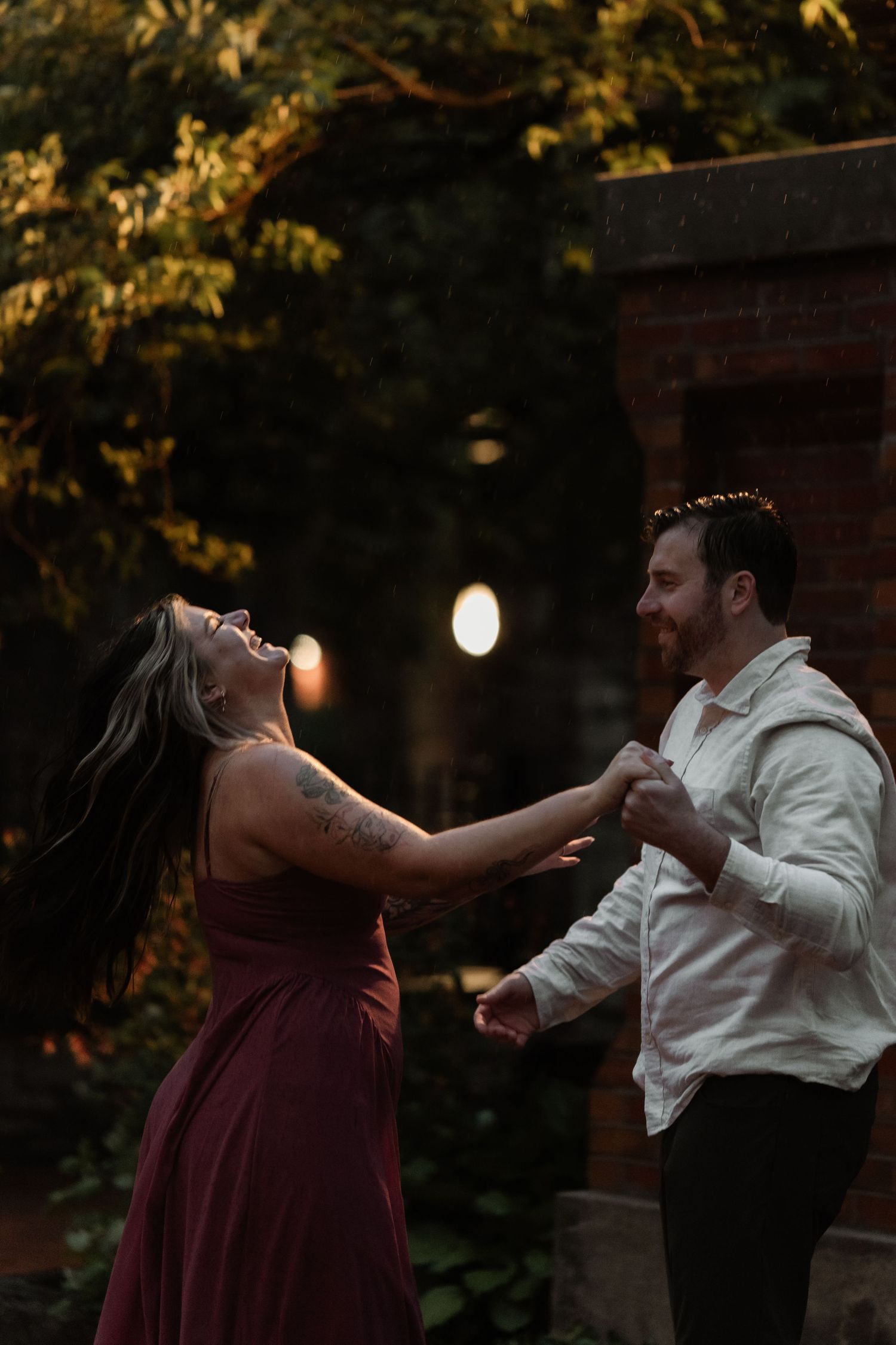 A joyful couple dances outdoors at dusk, woman in burgundy gown laughing, man in white shirt, surrounded by autumn foliage.