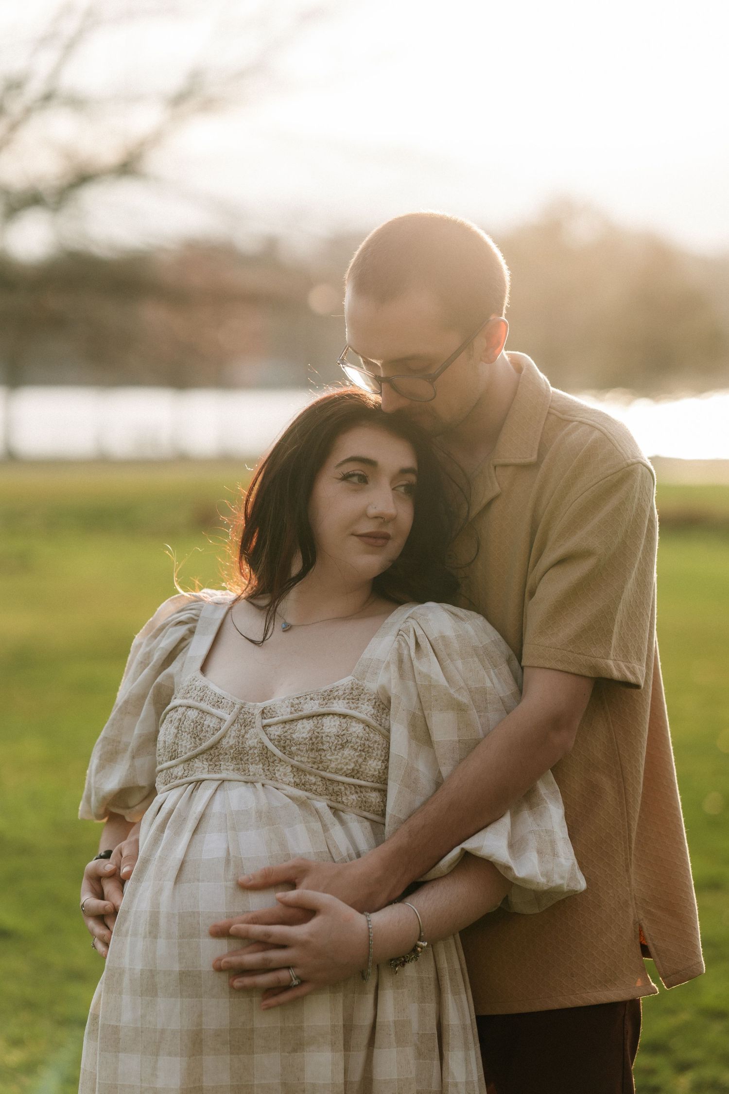Couple embracing outdoors at golden hour, pregnant woman in plaid dress with partner holding her belly in a sunlit park.