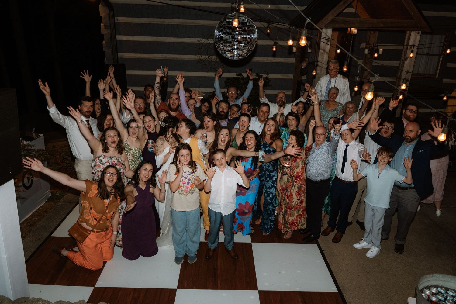 Large wedding party group photo on dance floor, guests cheering with arms raised under disco ball.