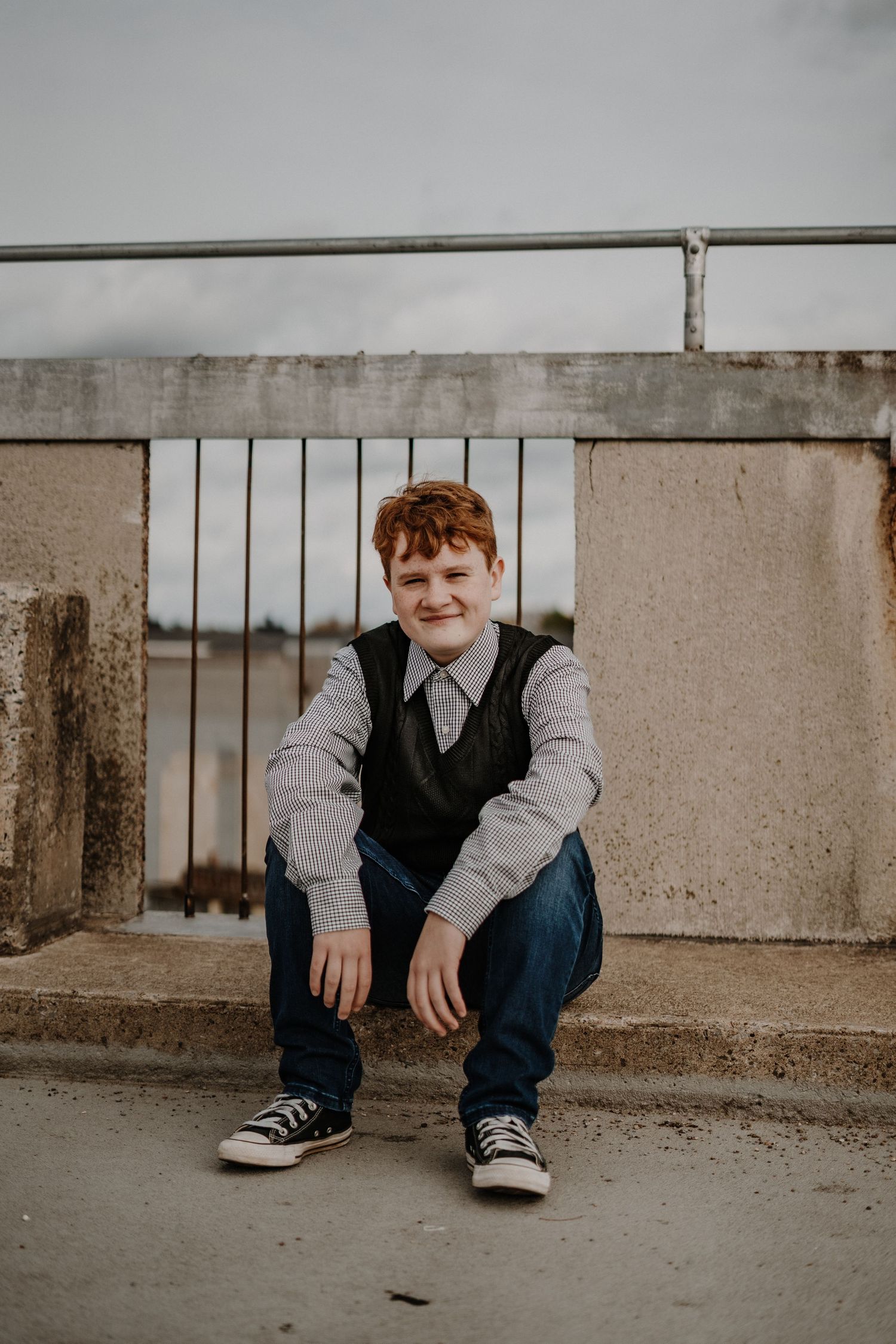 Senior portrait of person in vest and jeans sitting casually on a concrete ledge on an overcast day.