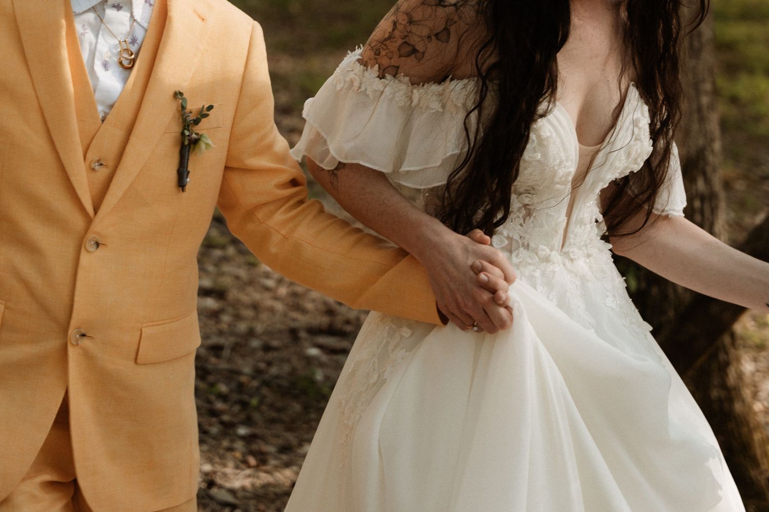 Bride in white lace gown holds hands with groom in yellow suit during outdoor wedding ceremony.