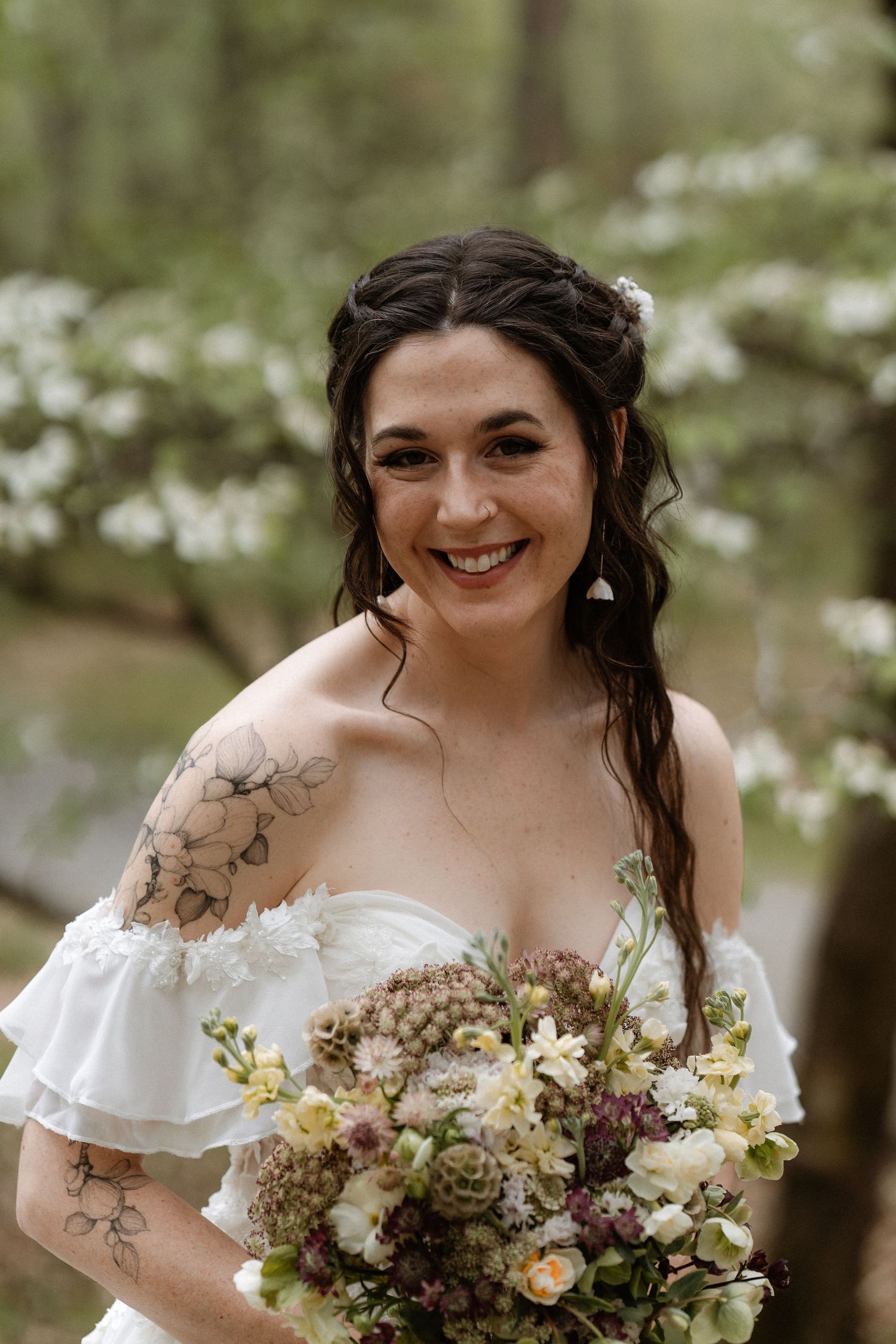 Smiling bride in off-shoulder white gown holds wildflower bouquet with succulents, posing before blooming trees.